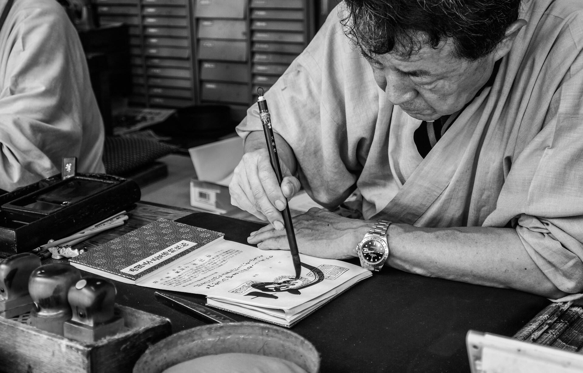 Fotografía en blanco y negro de un artesano que escribe con esmero en caligrafía con un pincel en un banco de trabajo, rodeado de papeles, herramientas y tinta, lo que crea una atmósfera de concentración.