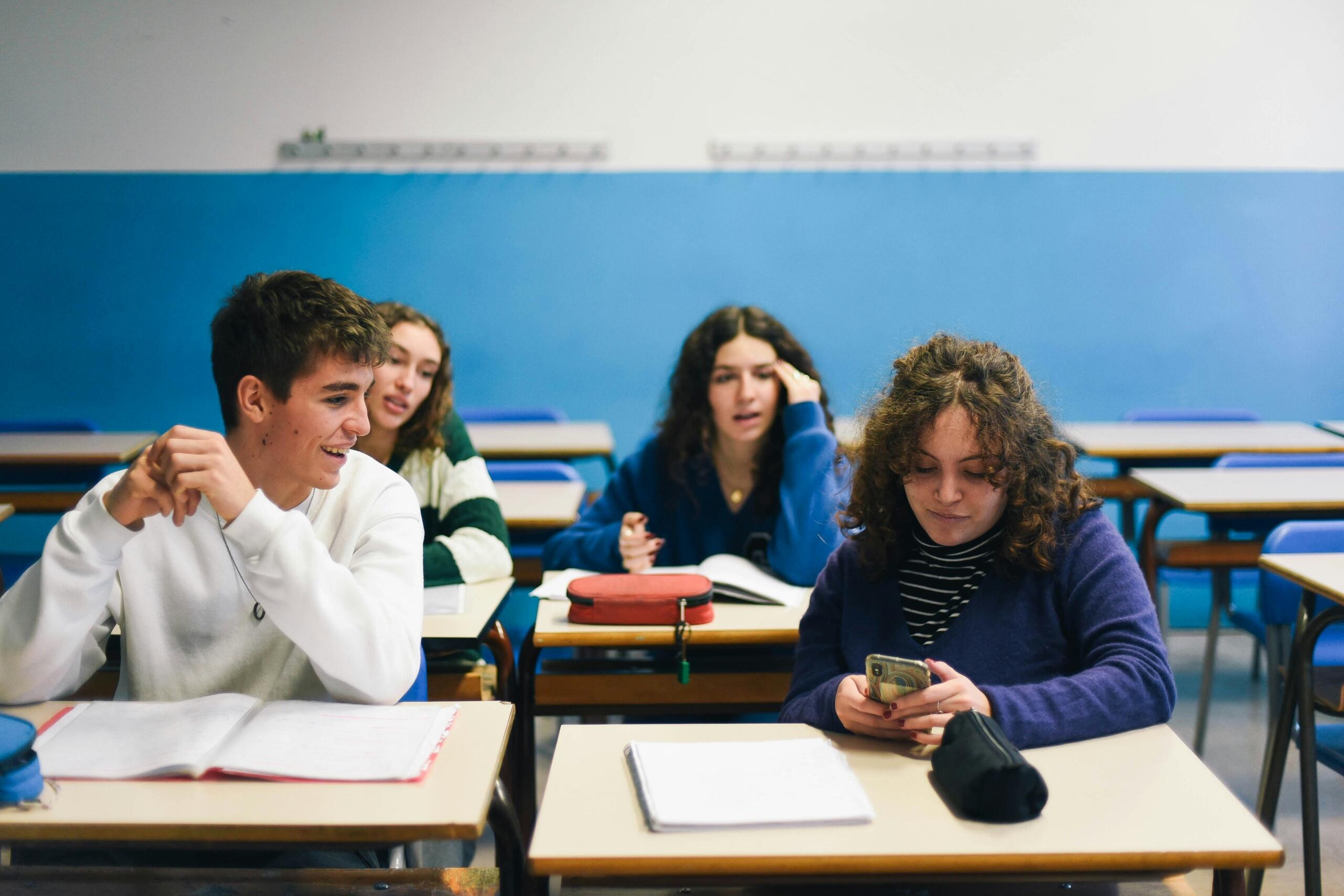 Los alumnos se sientan en los pupitres de un aula, ocupados con sus dispositivos y cuadernos, rodeados por una pared azul y sillas vacías.