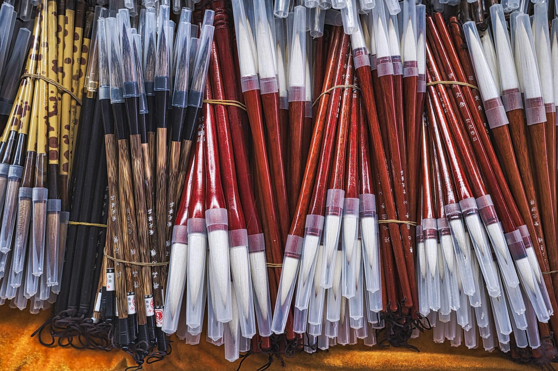 Un expositor está repleto de paquetes de pinceles de caligrafía china con mangos rojos, negros y amarillos, muchos de ellos protegidos por fundas transparentes; todo resulta muy colorido y ordenado.