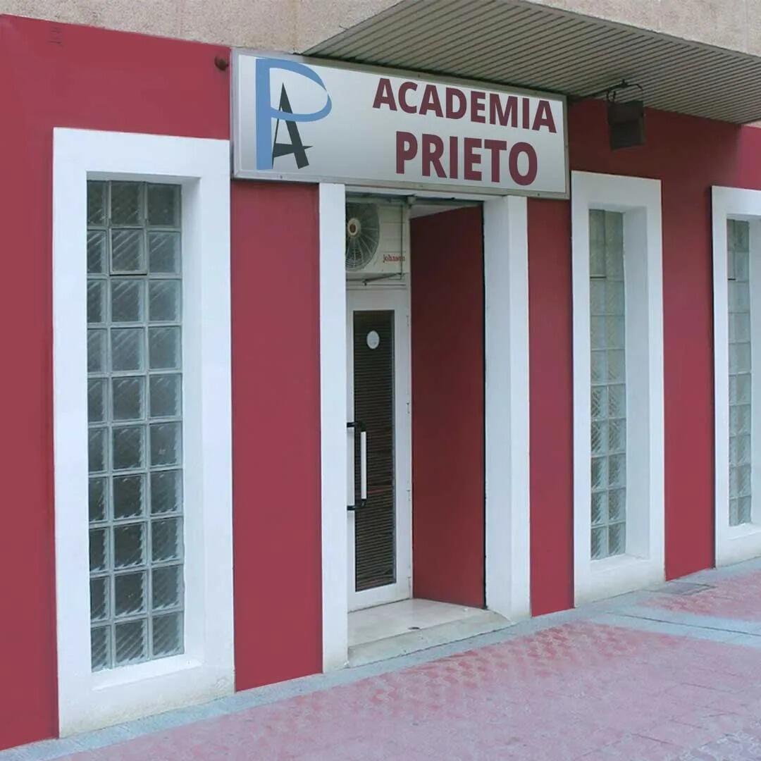 Edificio exterior rojo y blanco con un letrero que dice «Academia Prieto» sobre la puerta, flanqueado por ventanas verticales de ladrillo de vidrio.