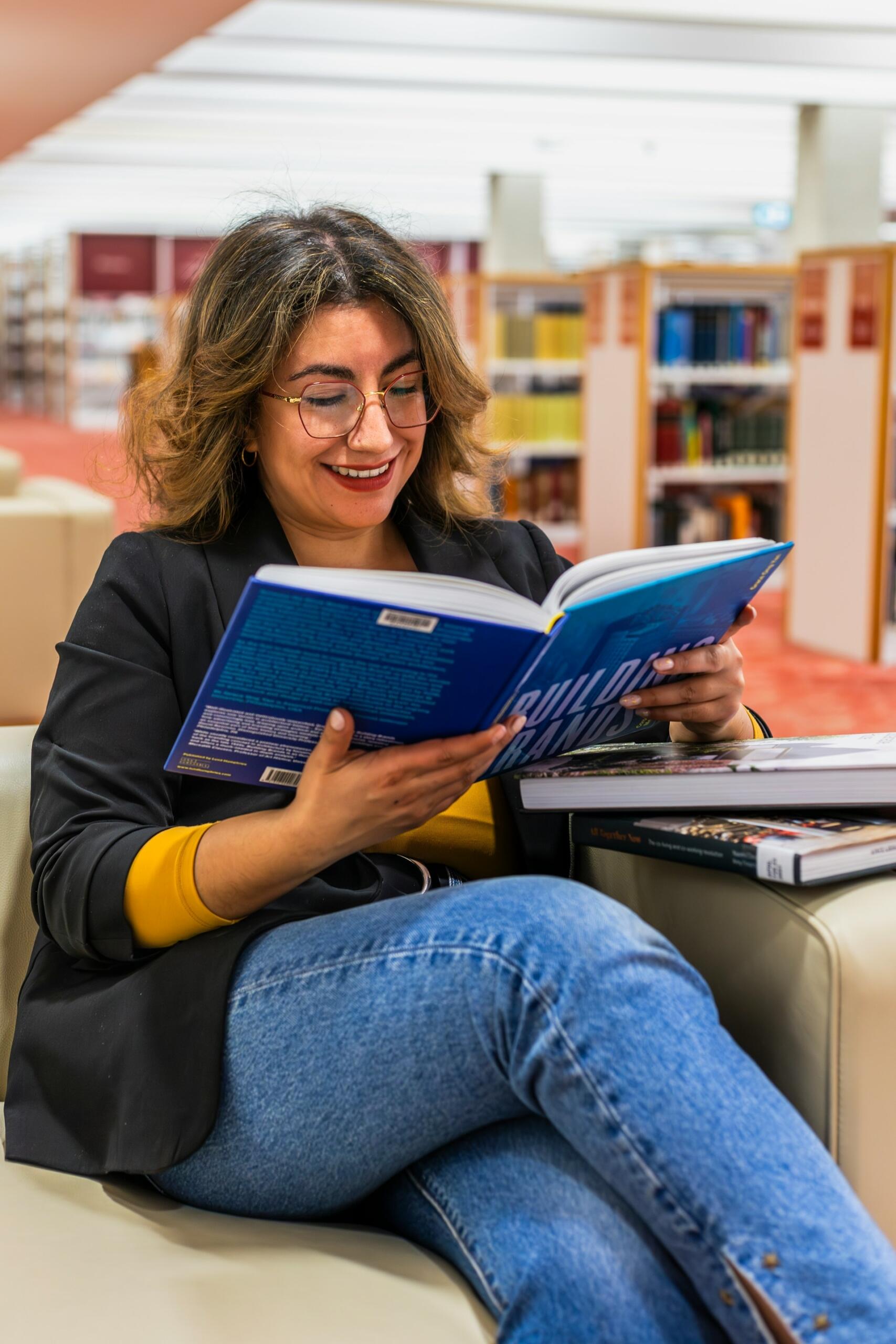 Una persona se sienta en un sofá de una biblioteca, leyendo un libro titulado "Construyendo marcas", rodeada de estantes de libros.