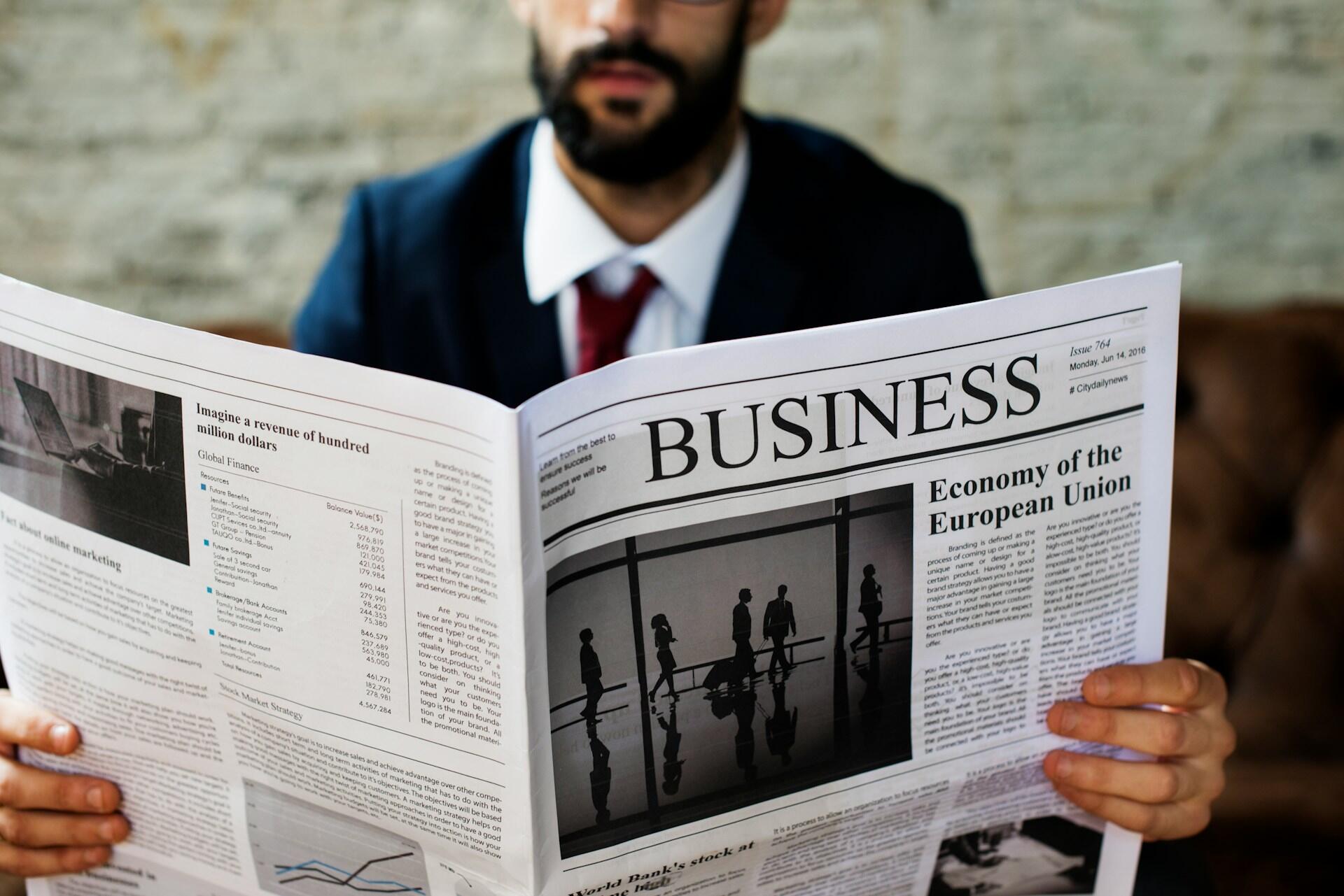 Una persona con traje sostiene un periódico económico con titulares sobre la economía y las finanzas mundiales, con un fondo borroso.