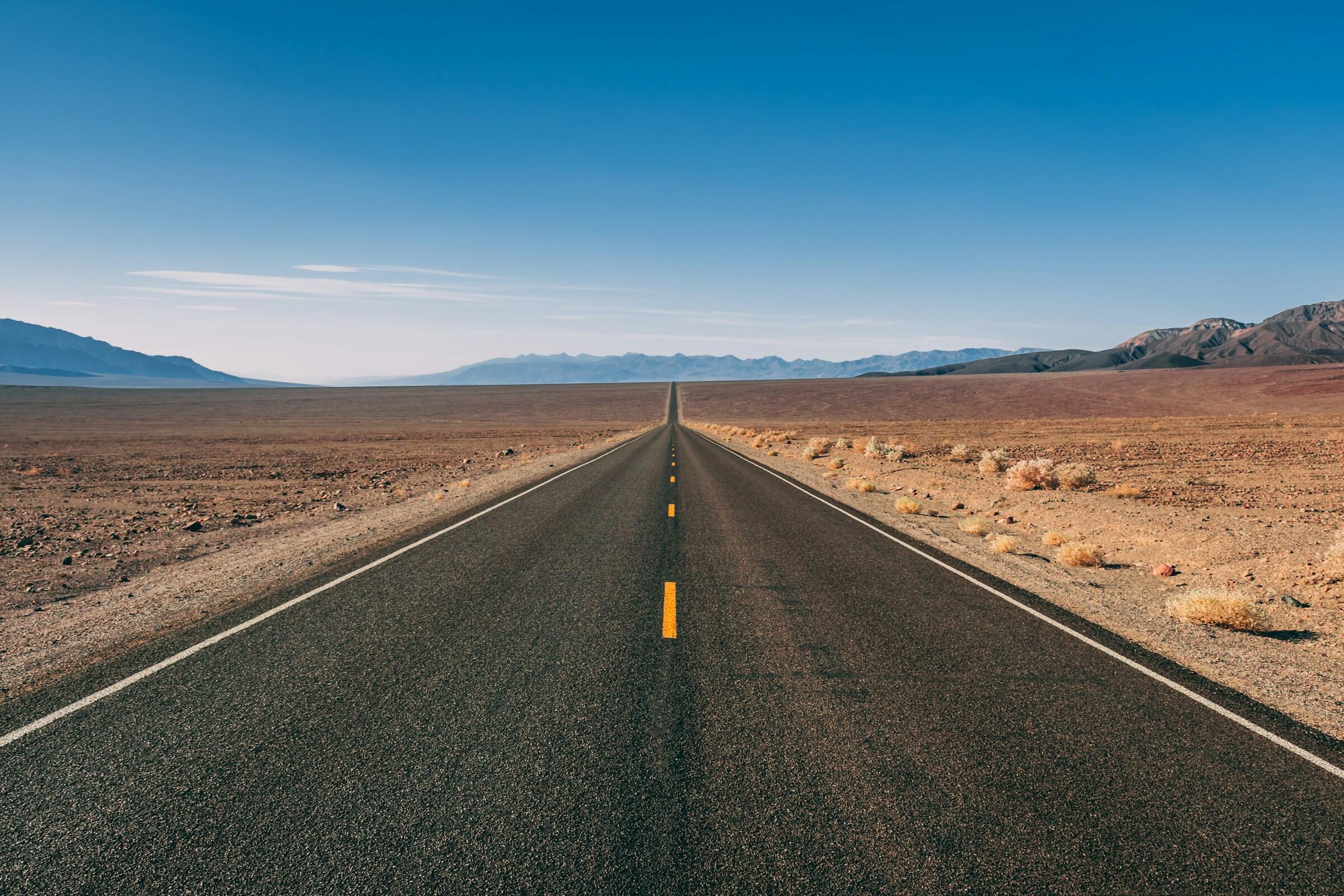 Una carretera larga y recta se extiende a través de un vasto y árido paisaje bajo un cielo azul claro y montañas distantes.