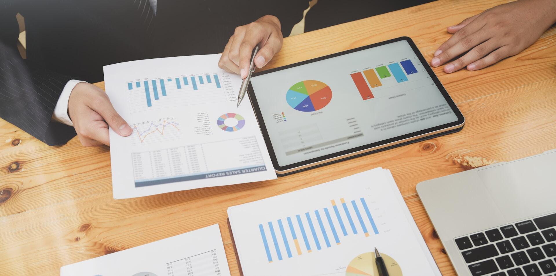 Dos manos analizan varios gráficos y cuadros en informes impresos y una tableta, mostrando el análisis de datos en un entorno empresarial.