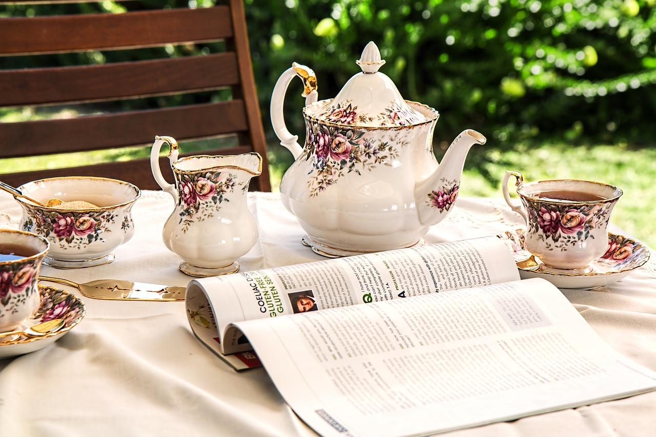 Elegante juego de té con diseños florales, azucarero y tetera sobre una mesa, junto a una revista abierta al aire libre.