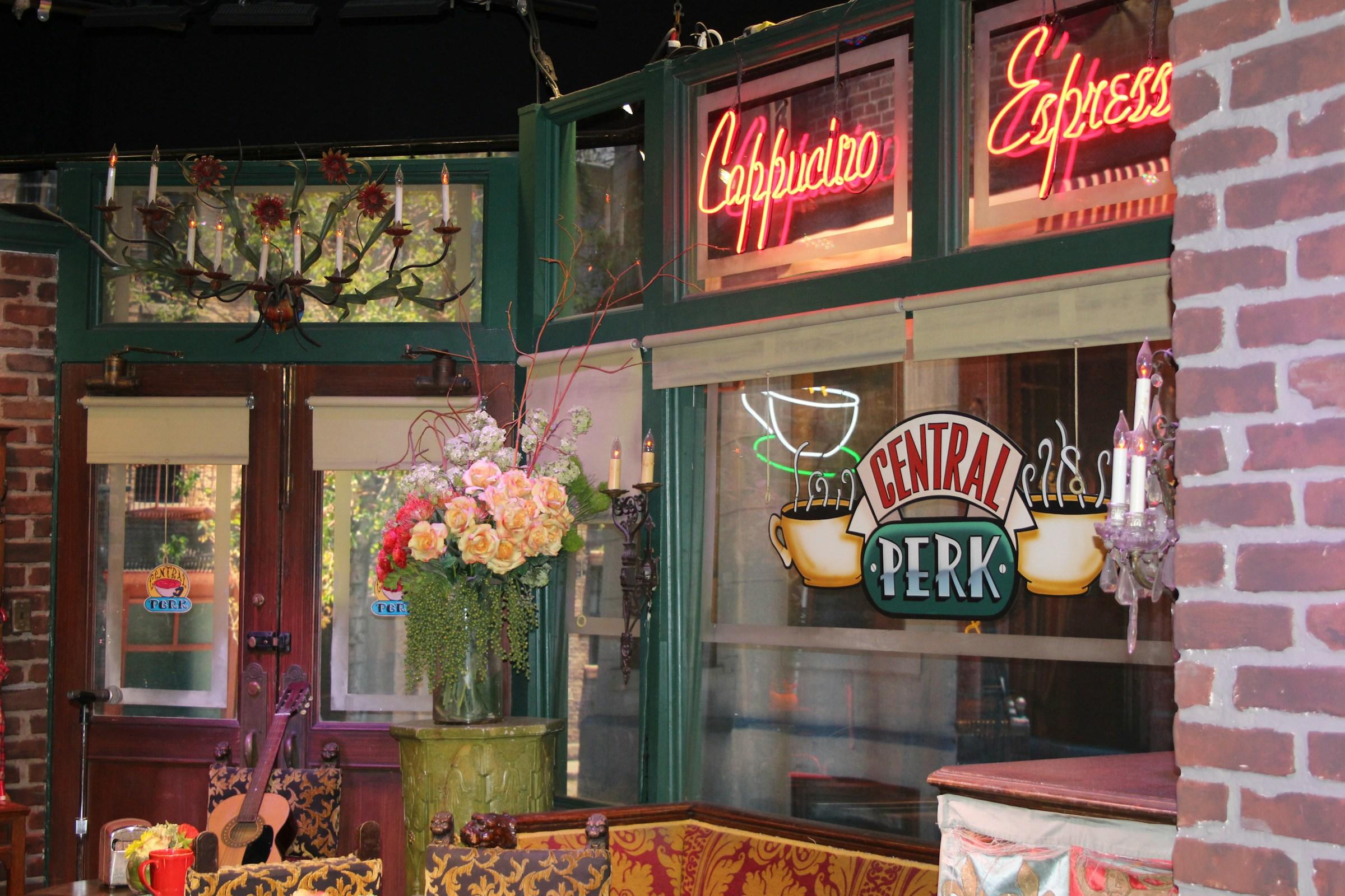 Acogedor interior de cafetería de la serie friends con carteles de neón 'Cappuccino Espresso', un gran arreglo floral y una guitarra.