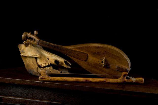 Un instrumento de cuerda rústico yace sobre una superficie de madera, con un cráneo de caballo a su lado. La composición transmite un tono sombrío e histórico.