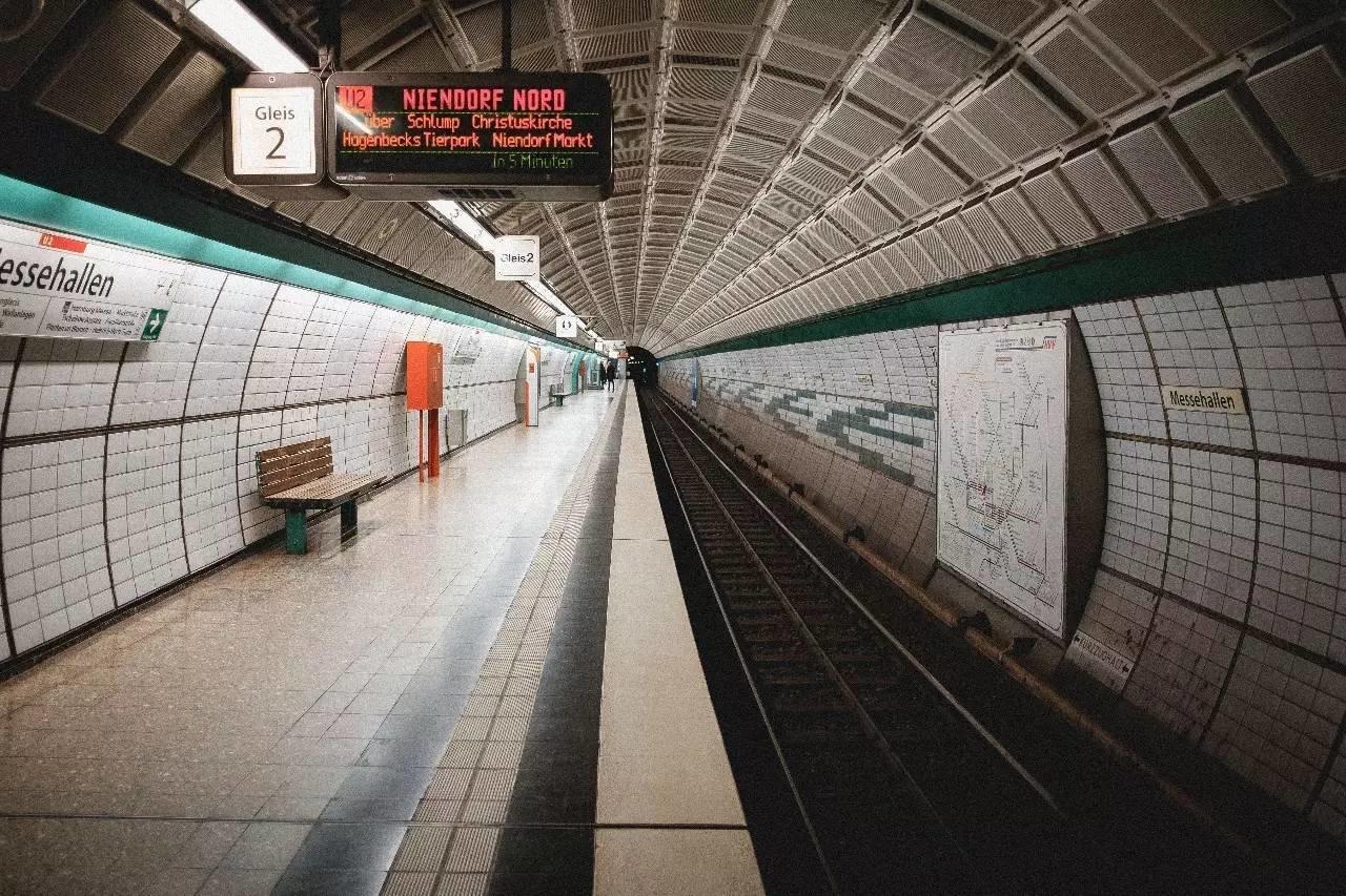 Un andén del metro de Berlín con azulejos blancos, una pantalla digital que indica la llegada de los trenes y un banco, con vías que conducen a un túnel.