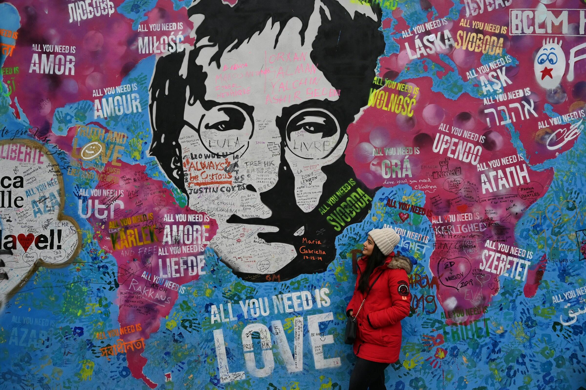 John Lennon con una chaqueta roja y un gorro blanco posa junto a un colorido mural en el que aparece el rostro de John Lennon y la frase «All you need is love» en varios idiomas.