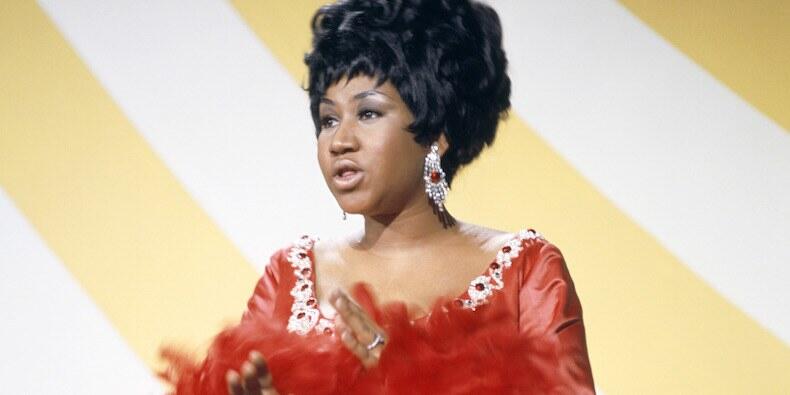 Aretha Franklin con un vestido rojo de mangas con plumas y pendientes grandes, cantando o hablando sobre un fondo a rayas amarillas y blancas.