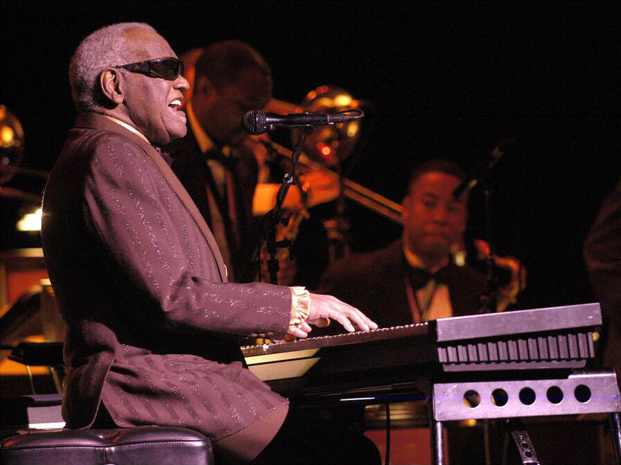 Ray Charles con gafas de sol toca el teclado mientras canta en el escenario, con los miembros de la banda al fondo.