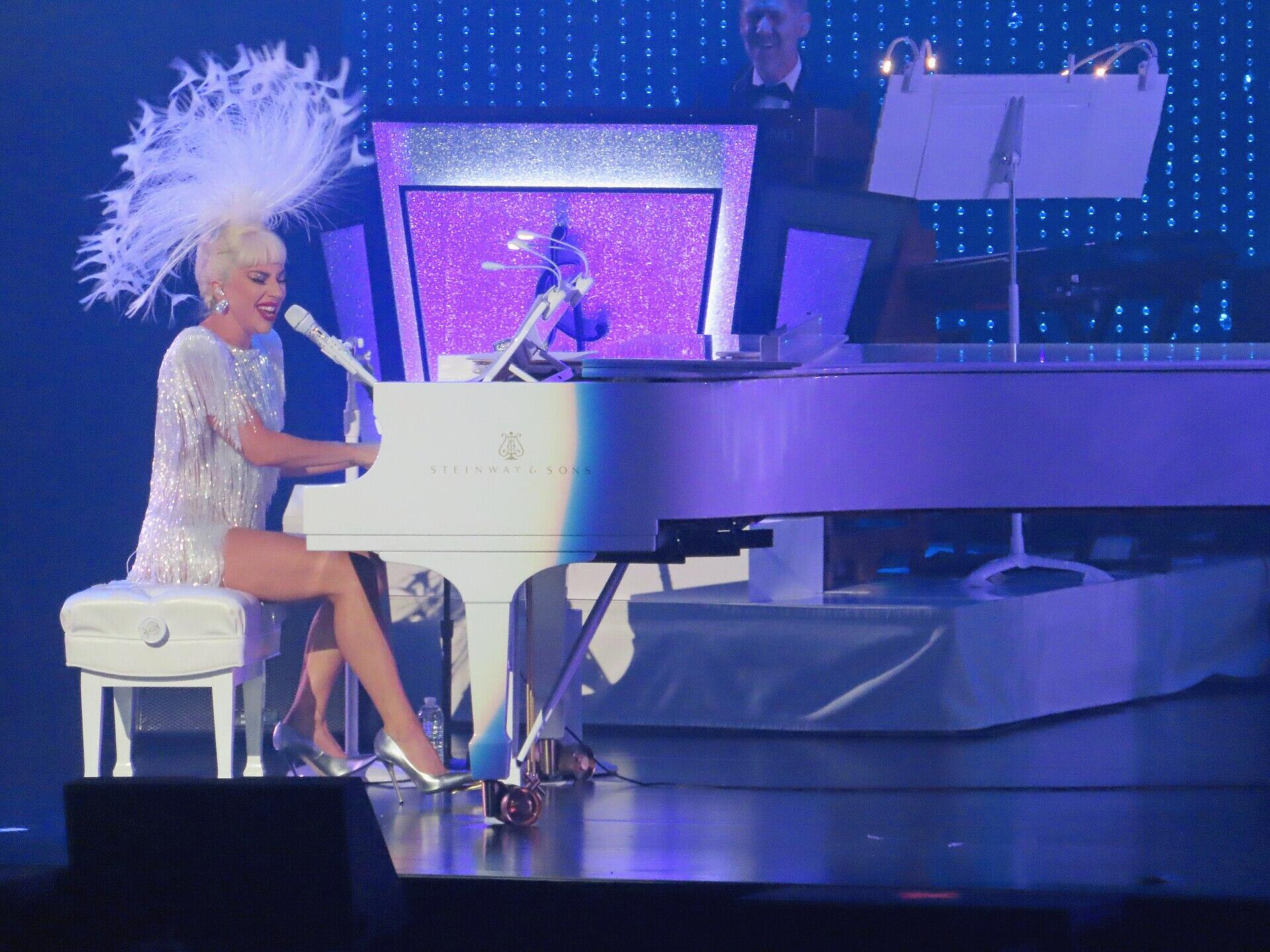 Lady gaga con un traje brillante y un tocado de plumas, toca un piano de cola blanco en el escenario, bajo una iluminación azul y morada.