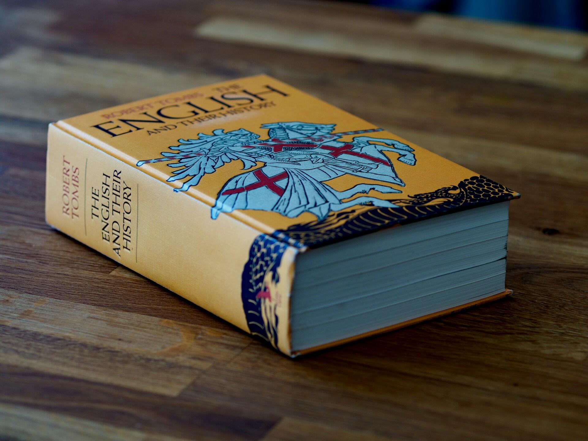 Libro de tapa dura amarillento y desgastado, «English and Their History», de Robert Tombs, con una ilustración de un caballero medieval, apoyado sobre una mesa de madera.