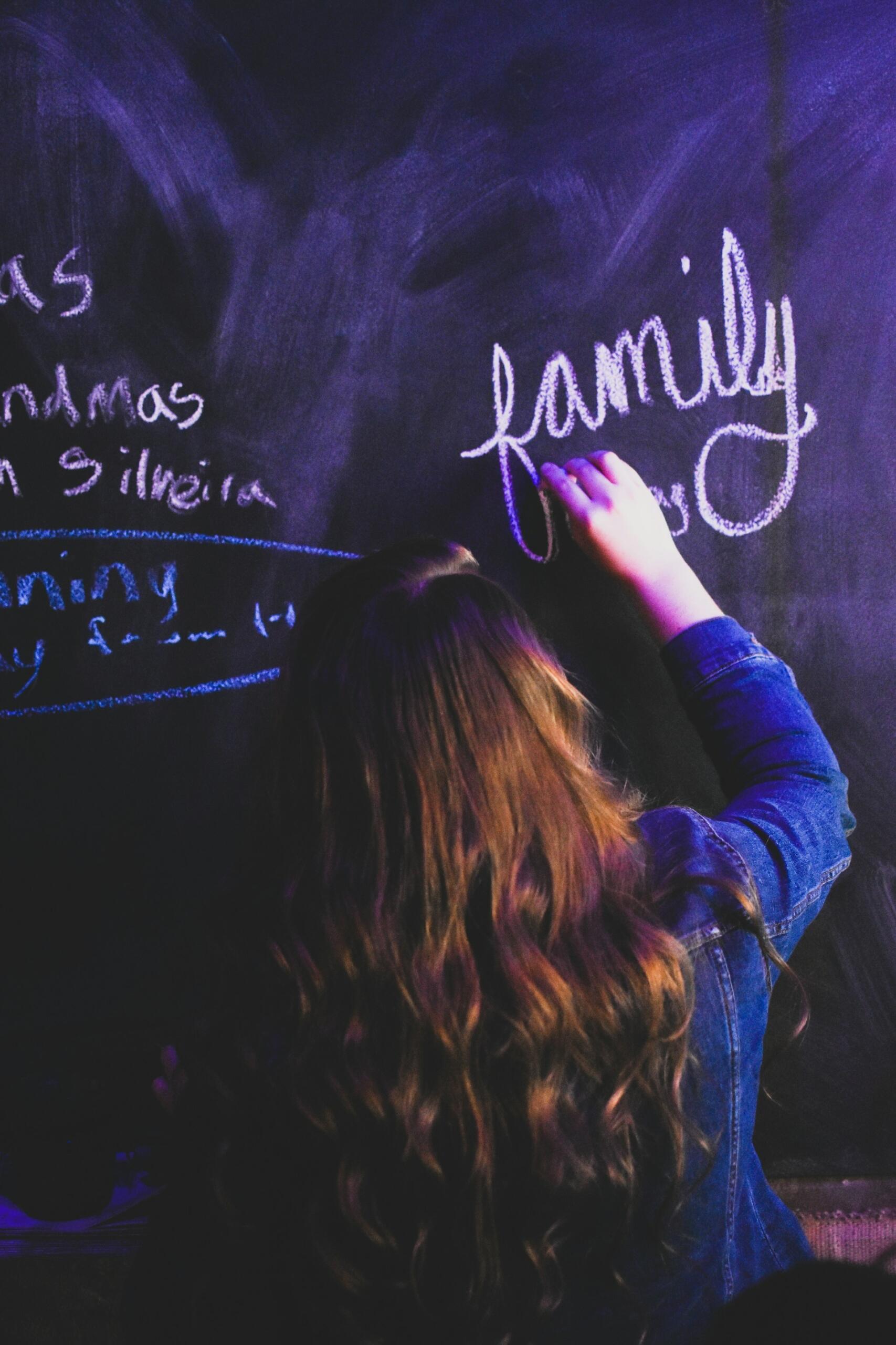 Una persona con el pelo largo y castaño escribe «familia» con tiza blanca en una pizarra oscura, iluminada por una luz morada; ambiente creativo y reflexivo.
