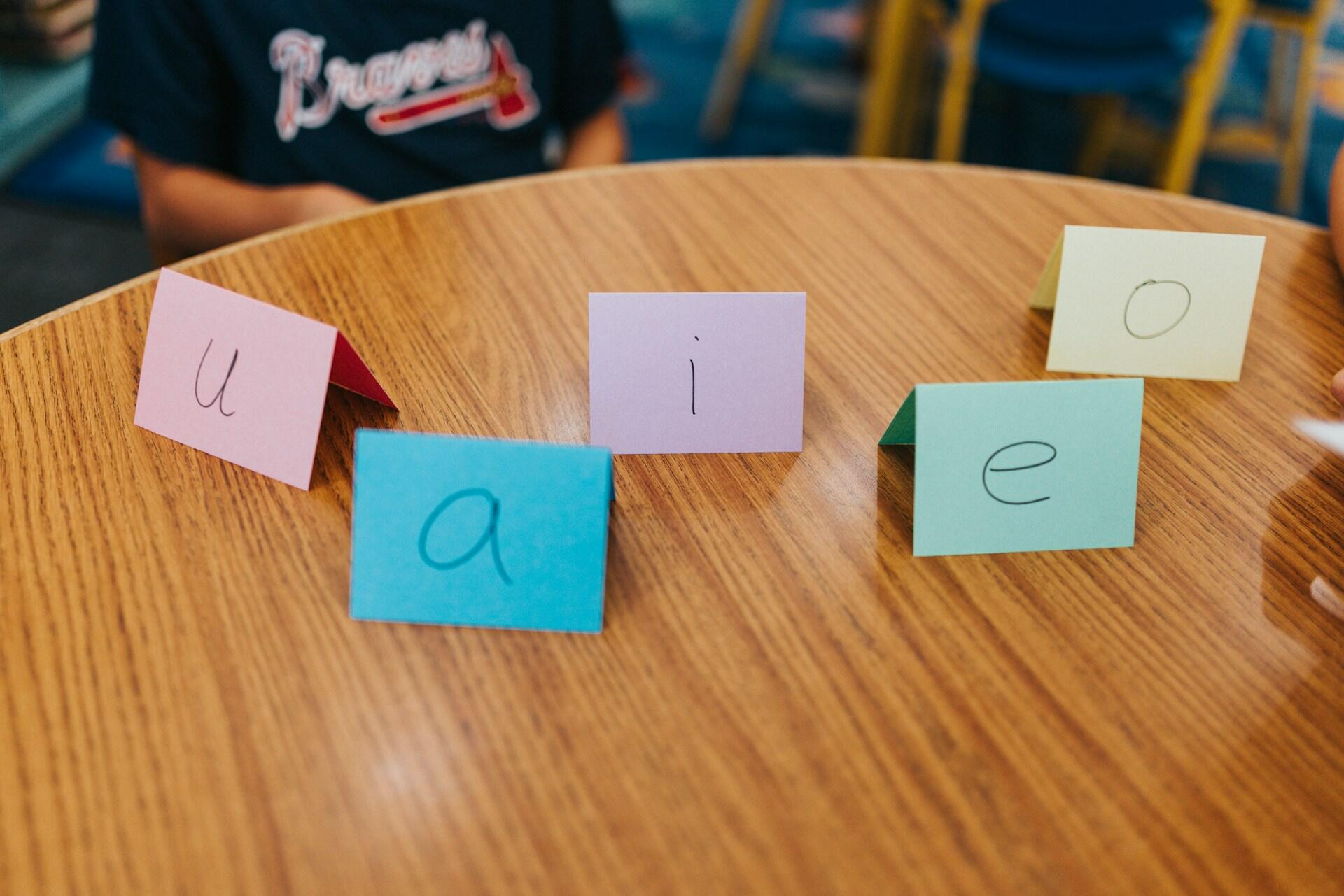 Fichas de colores con las vocales U, A, I, E y O escritas a mano, dispuestas sobre una mesa de madera, con un niño vestido con una camiseta deportiva detrás de ellas.