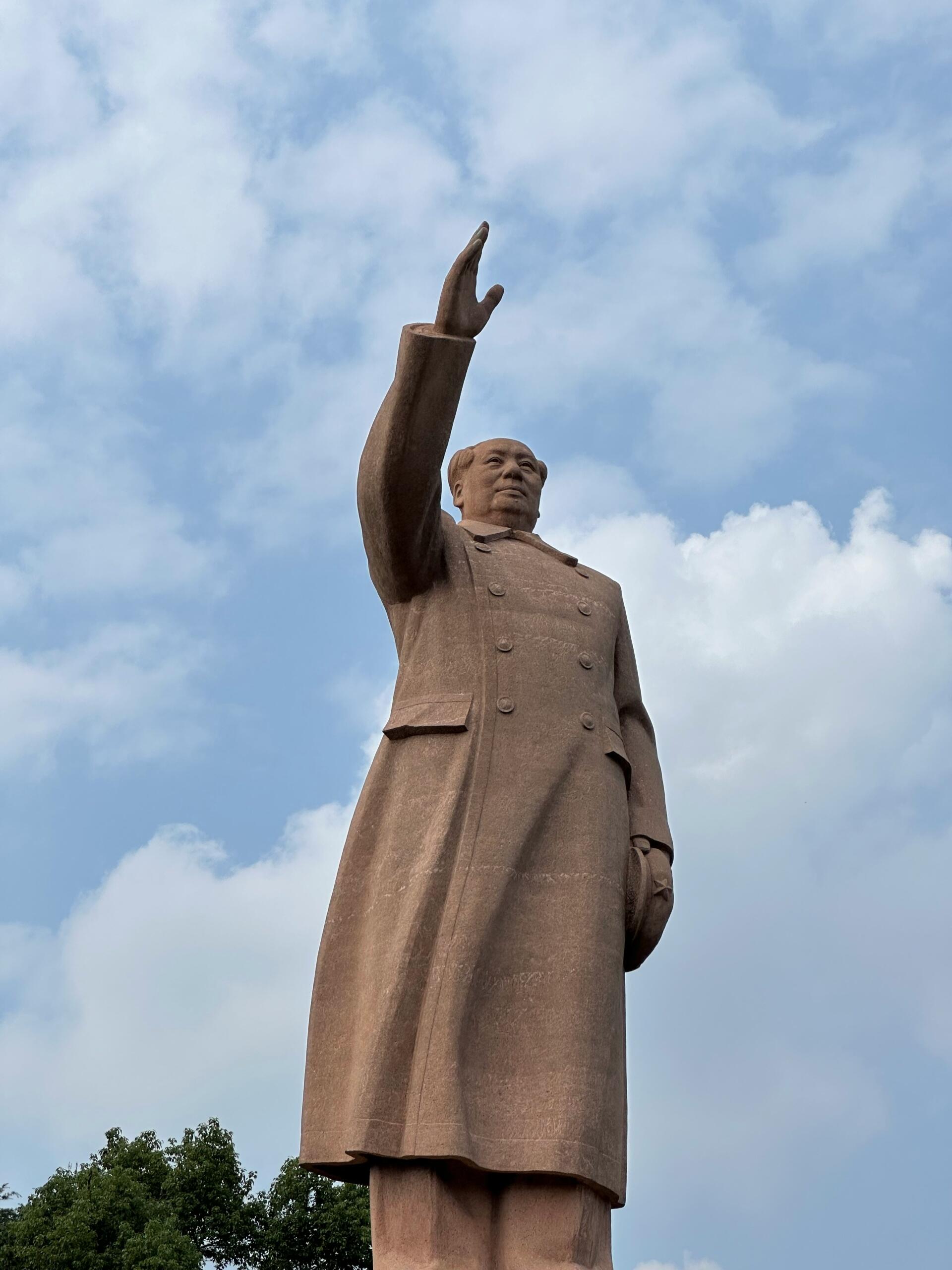 Estatua de Mao Zedong con un abrigo largo que levanta la mano derecha frente a un cielo parcialmente nublado, con árboles al fondo.
