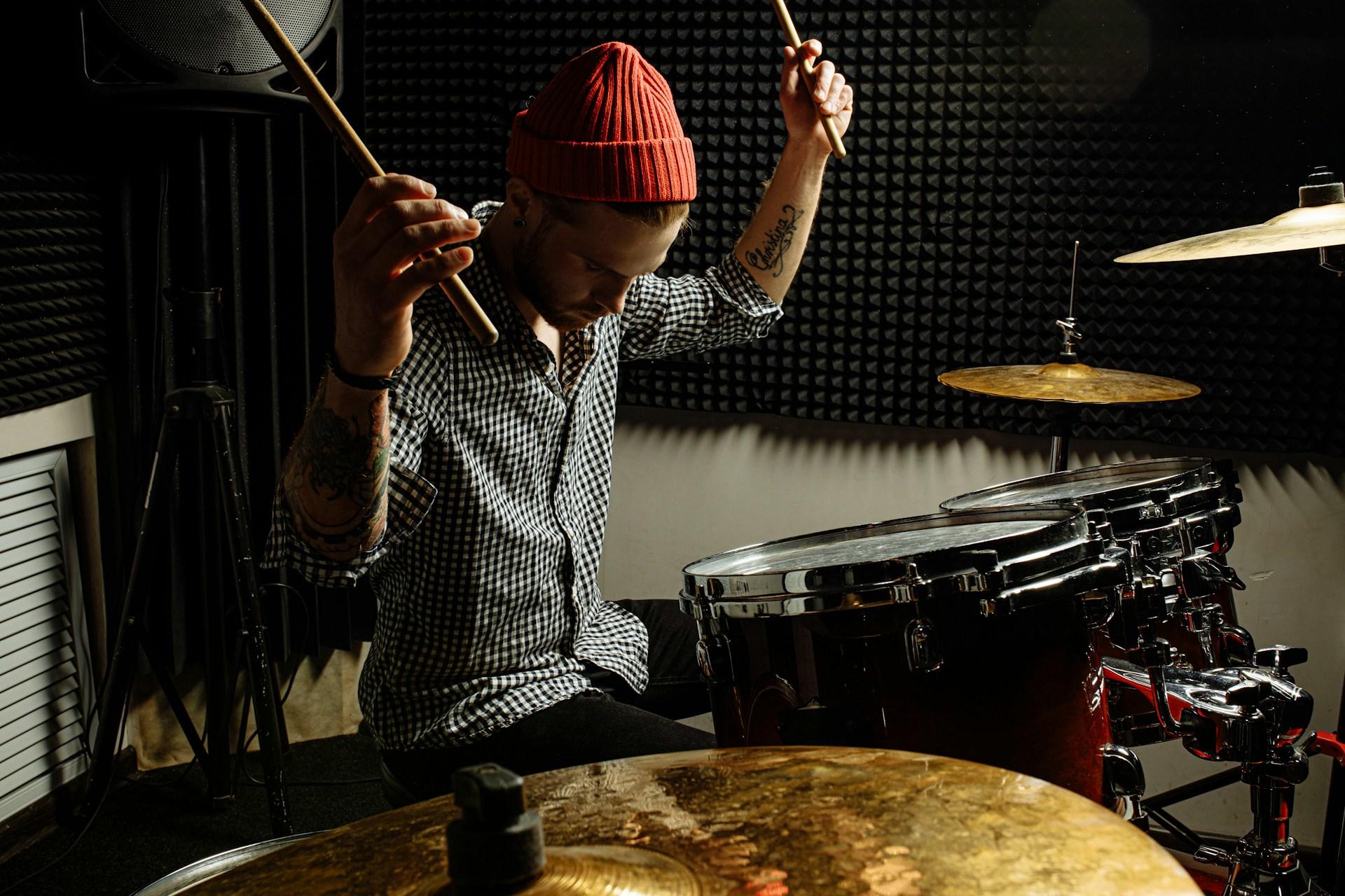 un hombre con gorro rojo y camisa gris tocando la batería en un estudio. Colores oscuros de la fotografía
