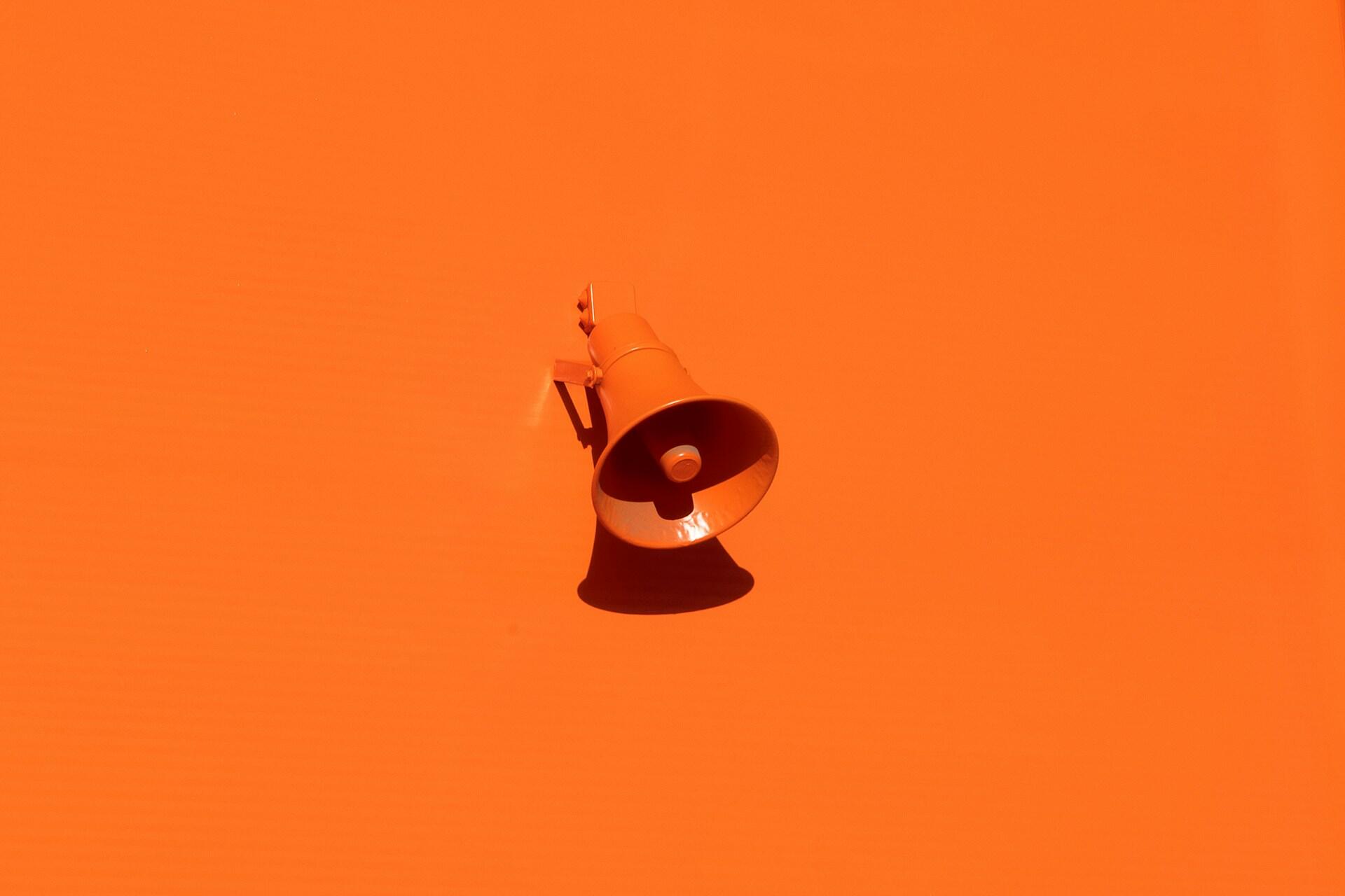 Un megáfono naranja colocado sobre una pared naranja a juego, que proyecta una sombra oscura. La composición minimalista transmite un tono atrevido y llamativo.