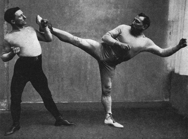 Dos hombres practican artes marciales tradicionales. Uno de ellos realiza una patada lateral y golpea el pecho del otro. Ambos muestran concentración e intensidad.