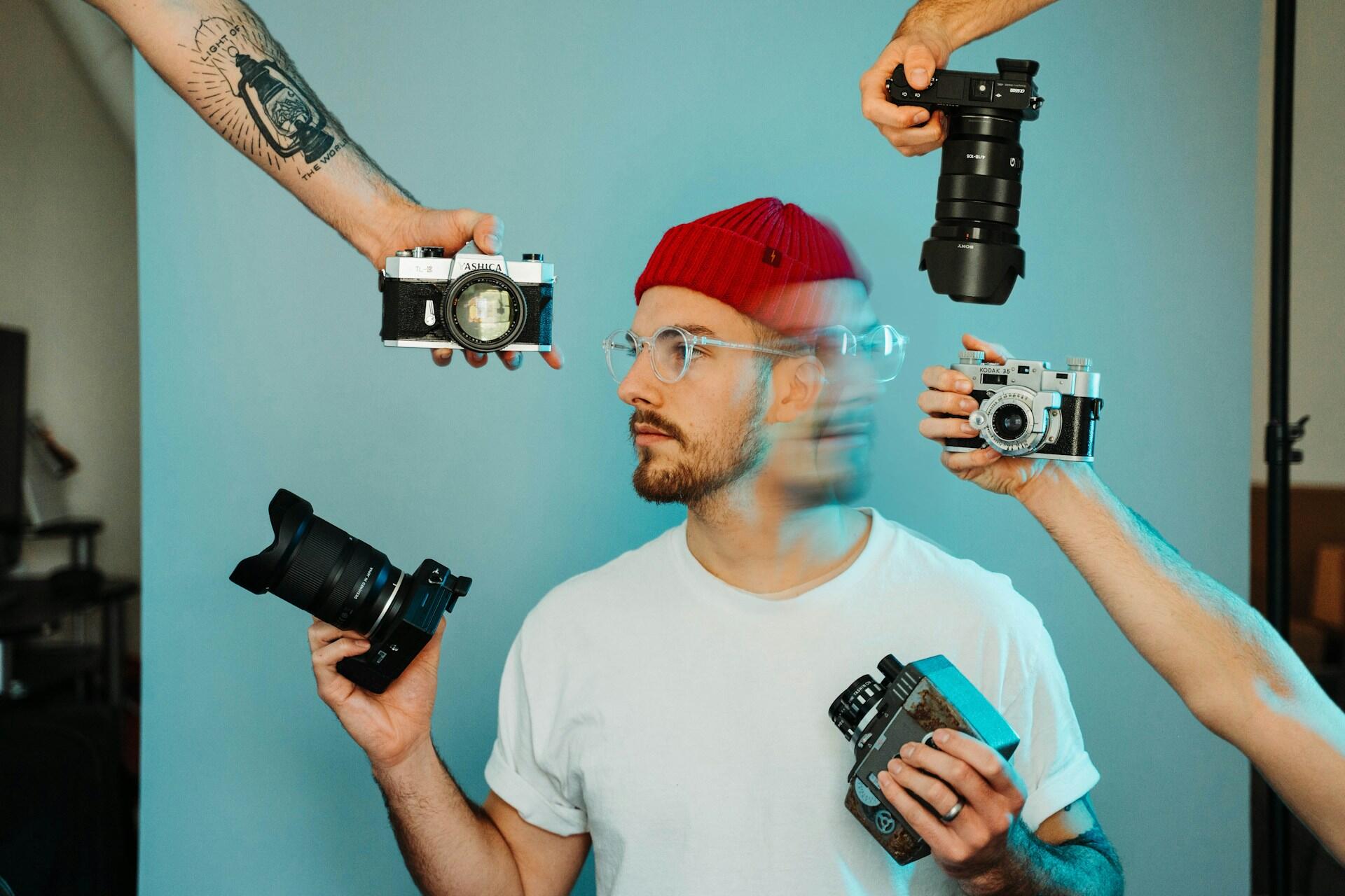Un hombre con un gorro rojo, una camiseta blanca y gafas está rodeado de manos que sostienen diversas cámaras sobre un fondo azul, lo que crea un ambiente divertido y curioso.