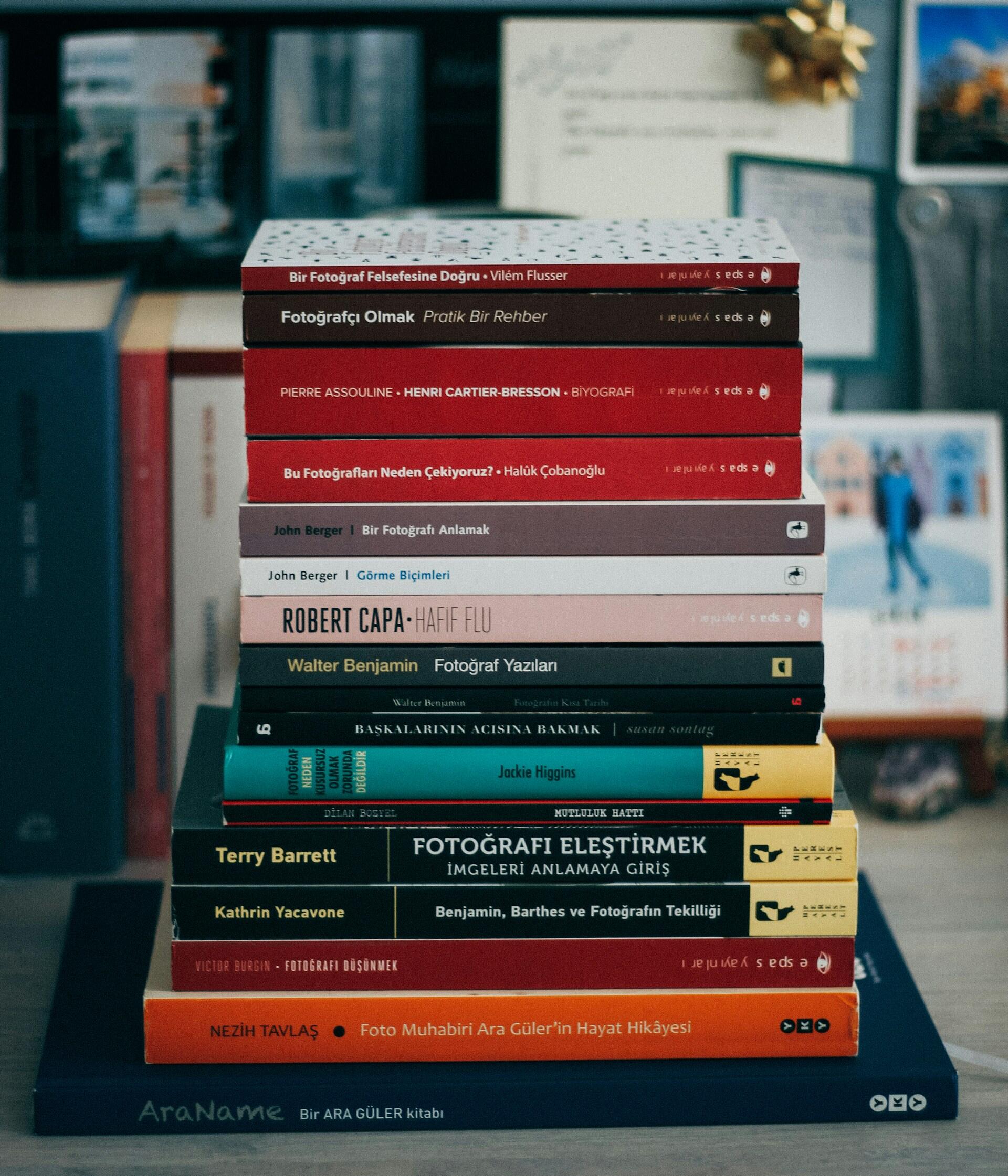 Sobre una mesa hay una pila de libros de fotografía con cubiertas de distintos colores. Al fondo se ven libros y papeles difuminados, junto con un pequeño adorno dorado, lo que transmite un ambiente erudito y artístico.