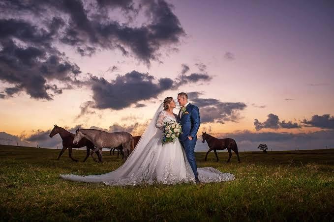 Una pareja vestida de boda posa cariñosamente en un prado al atardecer, con caballos pastando al fondo bajo un cielo nublado y espectacular.