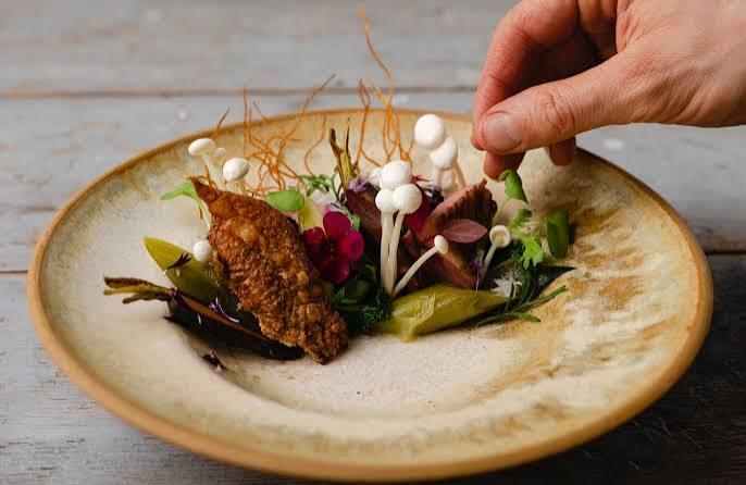 Un plato gourmet dispuesto a mano en un plato rústico con pollo crujiente, setas enoki, verduras de colores y flores comestibles, que transmite arte y elegancia.
