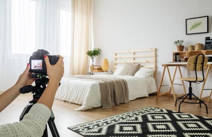 Se ha fotografiado un acogedor dormitorio con una cámara montada en un trípode. La habitación cuenta con una cama bien hecha, un escritorio de madera, plantas y una alfombra estampada, lo que crea un ambiente sereno.