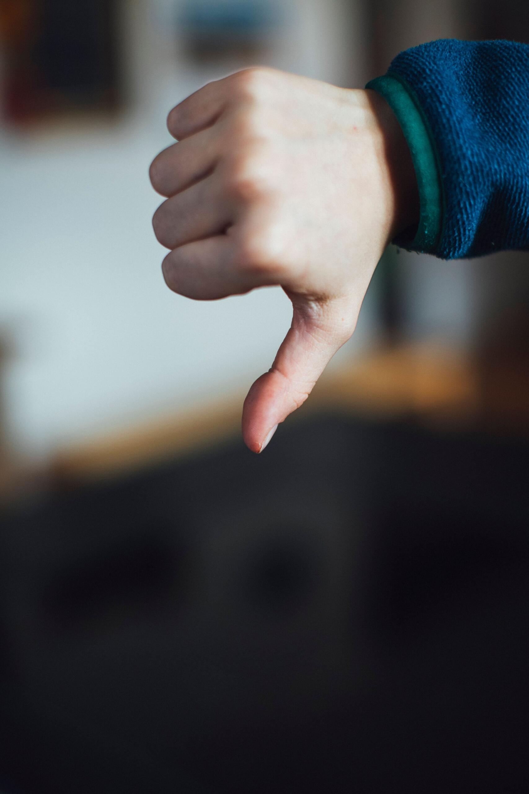 Primer plano de una mano haciendo el gesto del pulgar hacia abajo, que sobresale de una manga azul sobre un fondo ligeramente desenfocado, lo que transmite desaprobación.