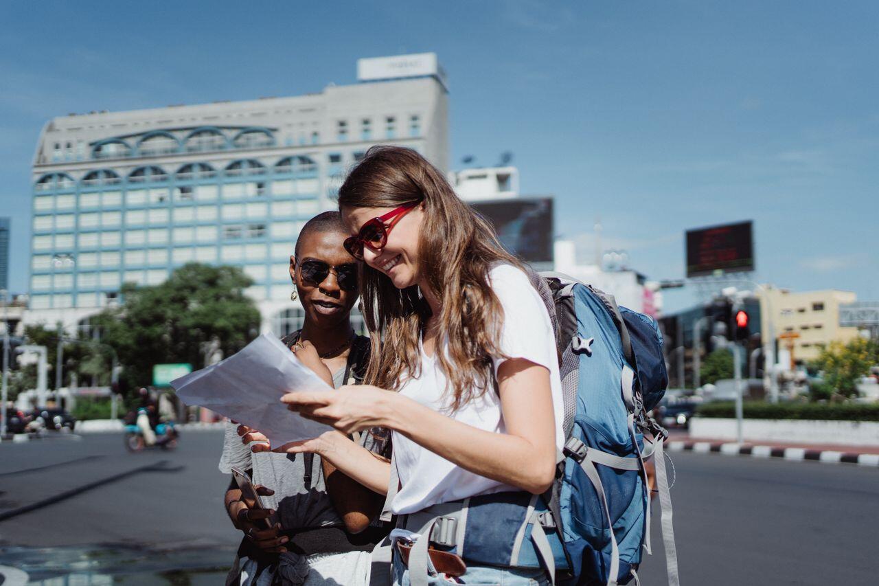 Dos viajeras examinan un mapa de papel en una calle soleada de la ciudad; una de ellas lleva gafas de sol y una mochila, lo que crea un ambiente alegre propio de un viaje.