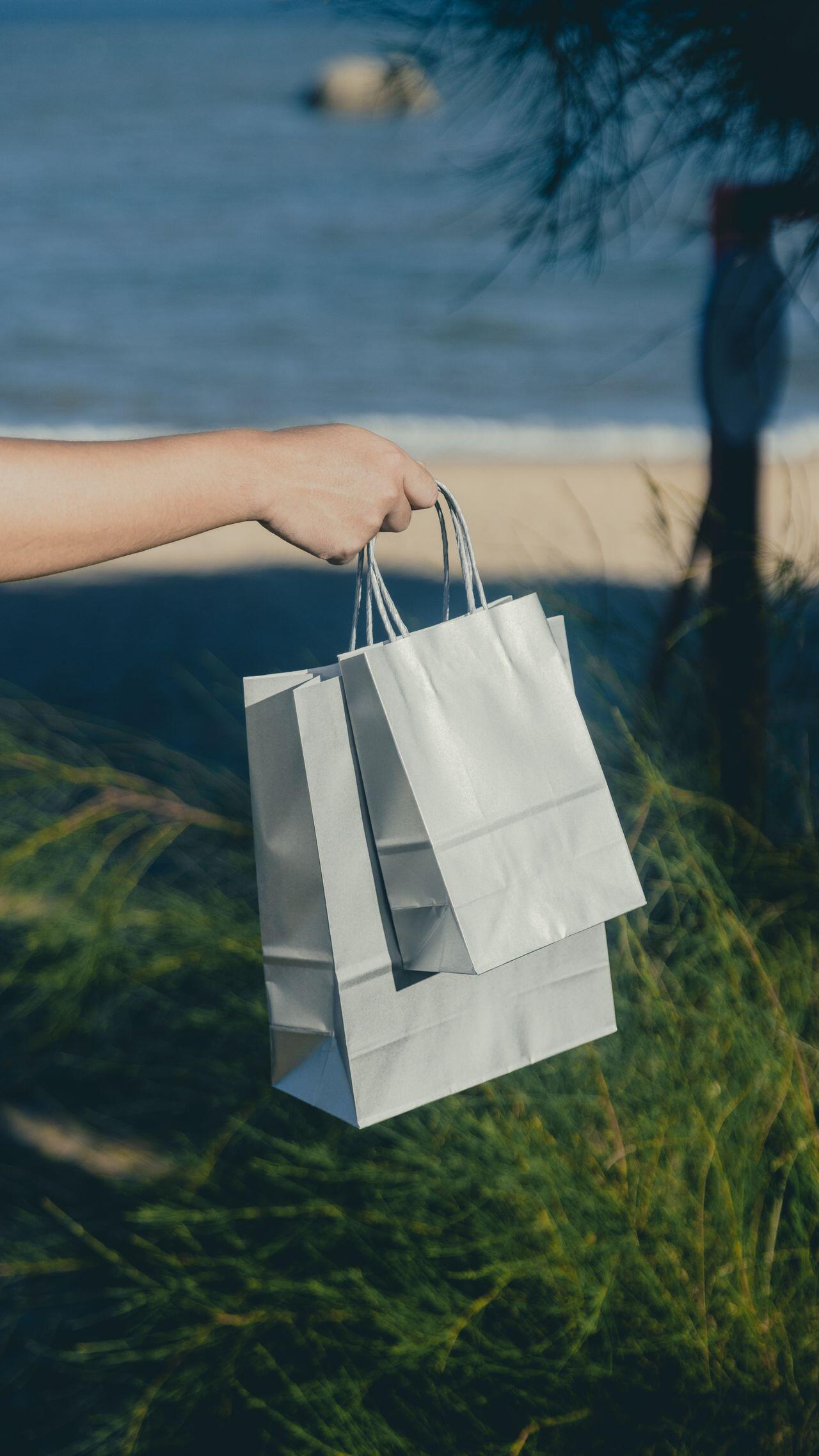 Una mano que sostiene unas bolsas de la compra de papel blanco cerca de una playa, con el mar, la arena, la hierba de las dunas y un bañista en la lejanía, creando un ambiente costero tranquilo.