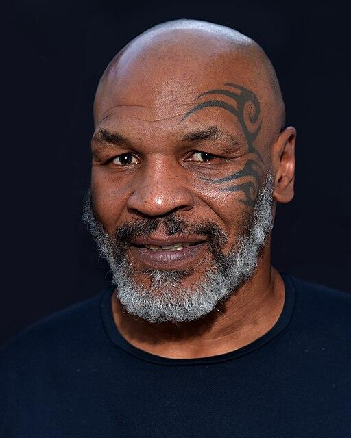 Mike Tyson con barba gris y un tatuaje facial en el lado izquierdo de la cara, vestido con una camisa negra, sonríe suavemente sobre un fondo oscuro.