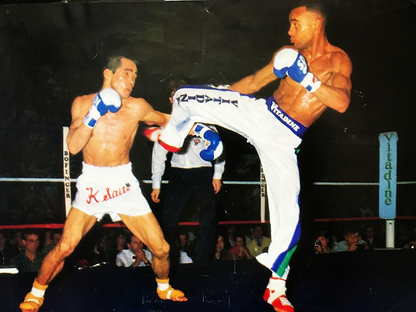 Dos kickboxers en un ring, uno lanzando una patada alta a la cara del otro. La escena es dinámica e intensa, con expresiones concentradas. Se ven espectadores al fondo.