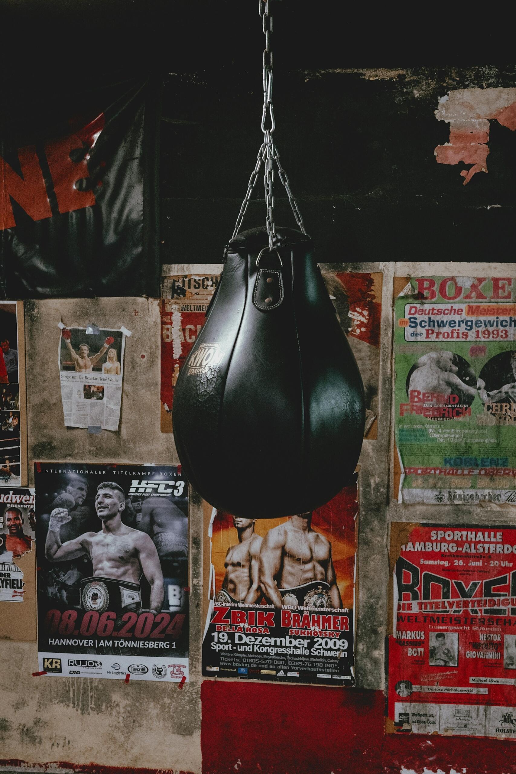 Un saco de boxeo negro cuelga en un viejo gimnasio con carteles de boxeo desgastados en una pared deteriorada. La escena transmite una atmósfera cruda y nostálgica.