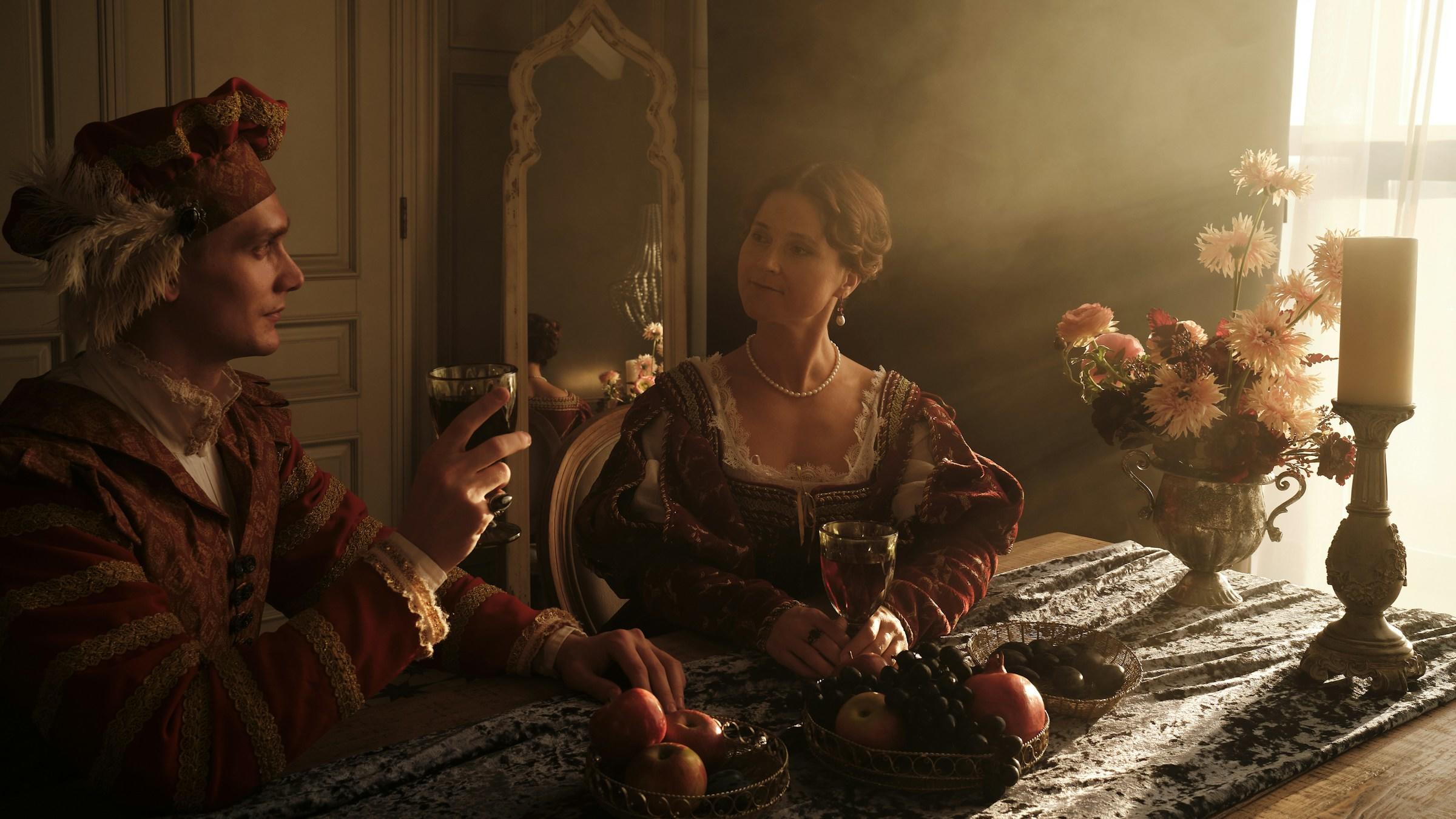 Dos individuos con atuendo histórico en una mesa con fruteros, velas y flores, en una habitación poco iluminada.