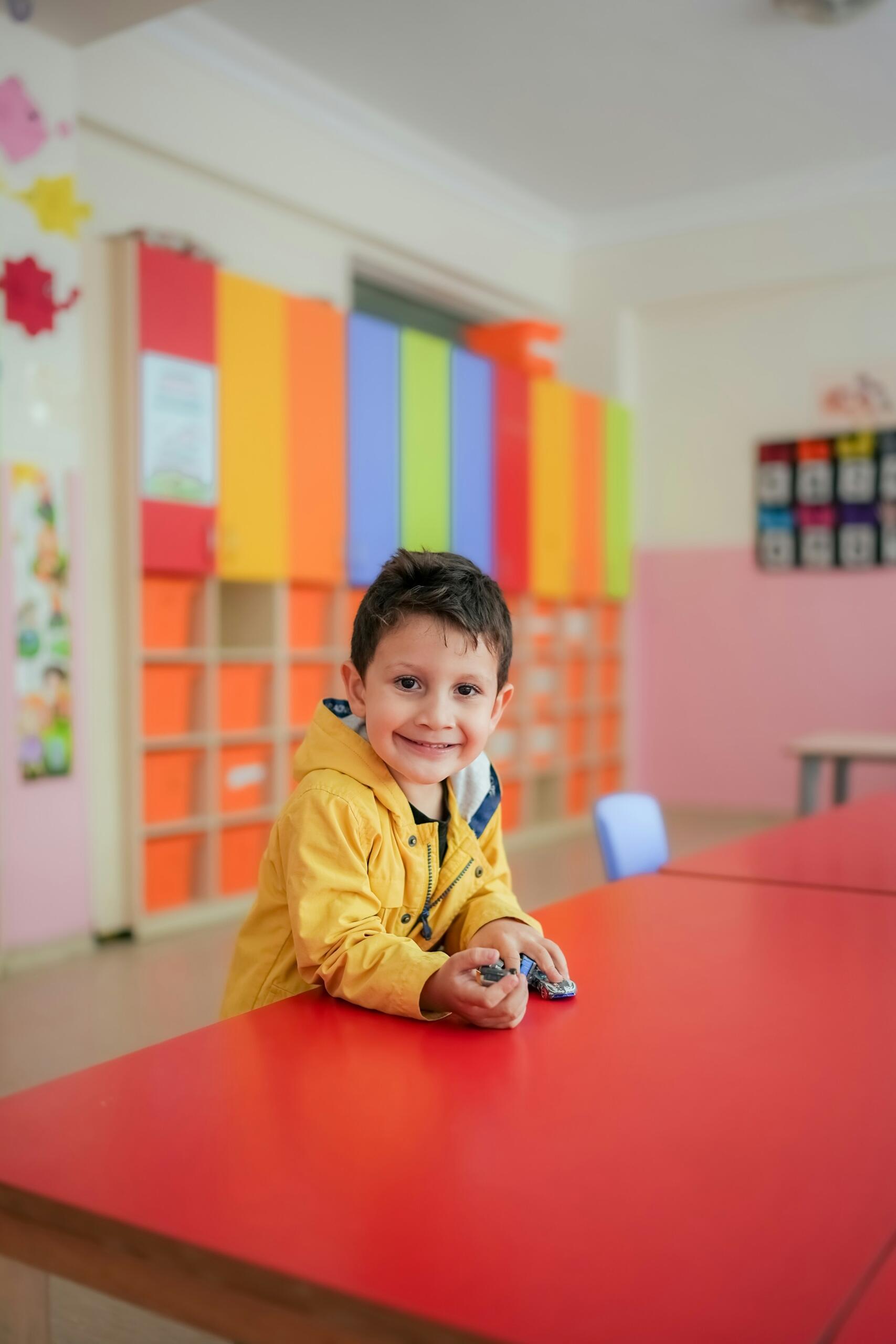 Un niño con una chaqueta amarilla está sentado en una mesa roja con unos coches de juguete en las manos, rodeado de coloridos armarios en un luminoso aula.
