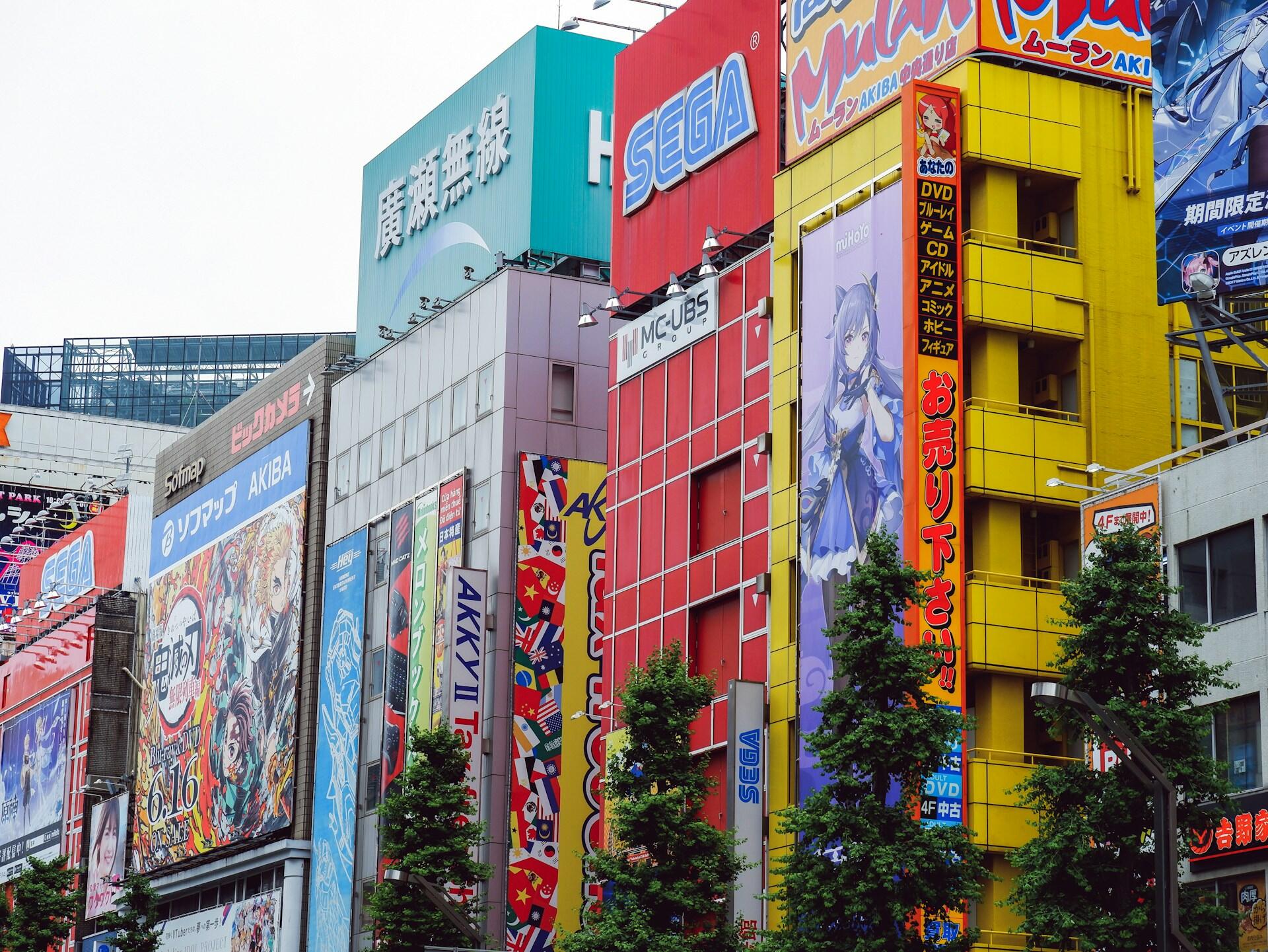 El colorido paisaje urbano de Akihabara, en Tokio, con vallas publicitarias de anime, letreros de SEGA y de tiendas de electrónica, tiendas altísimas, árboles y la bulliciosa energía urbana.