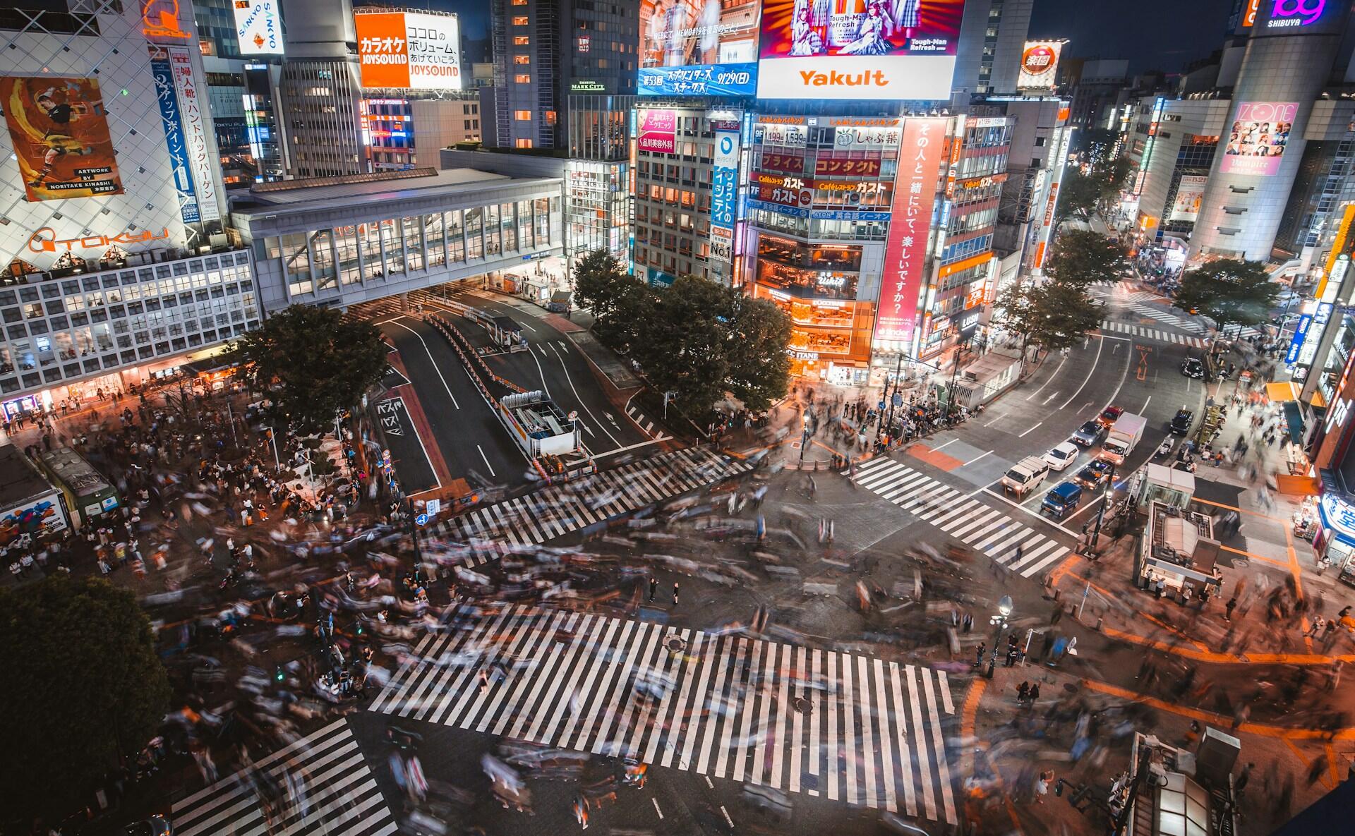 Vista aérea nocturna del cruce de Shibuya, en Tokio, repleto de peatones, tráfico y vallas publicitarias de neón que brillan en un animado paisaje urbano.
