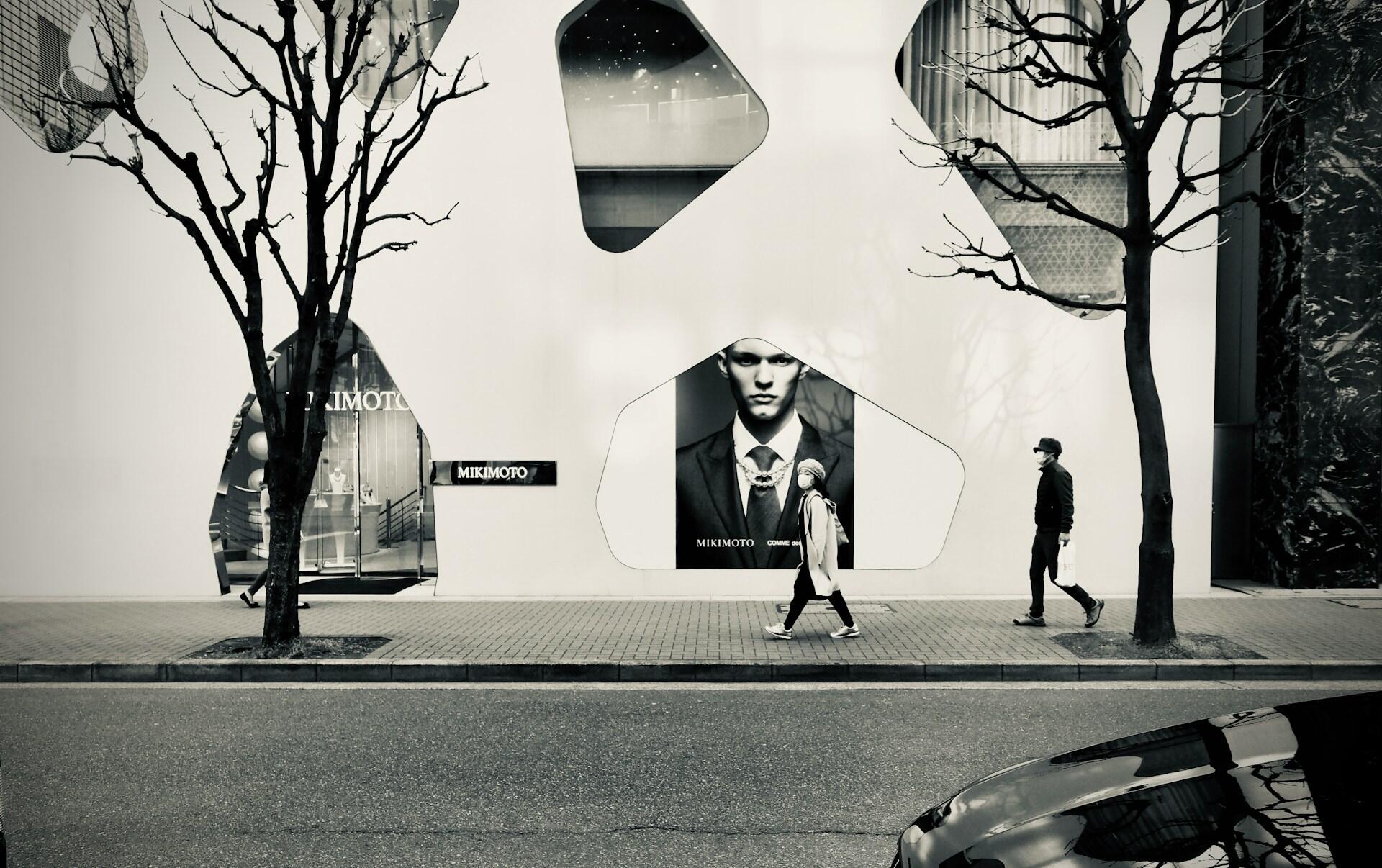 Calle urbana en blanco y negro con árboles sin hojas, peatones, escaparates modernos, un modelo vestido con traje en una valla publicitaria y un coche; tono evocador y cinematográfico.