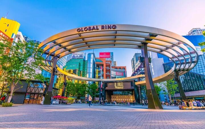 Una soleada plaza urbana bajo el «Global Ring», una marquesina metálica curvada, con rascacielos de colores, árboles, tiendas y peatones que crean un animado paisaje urbano.