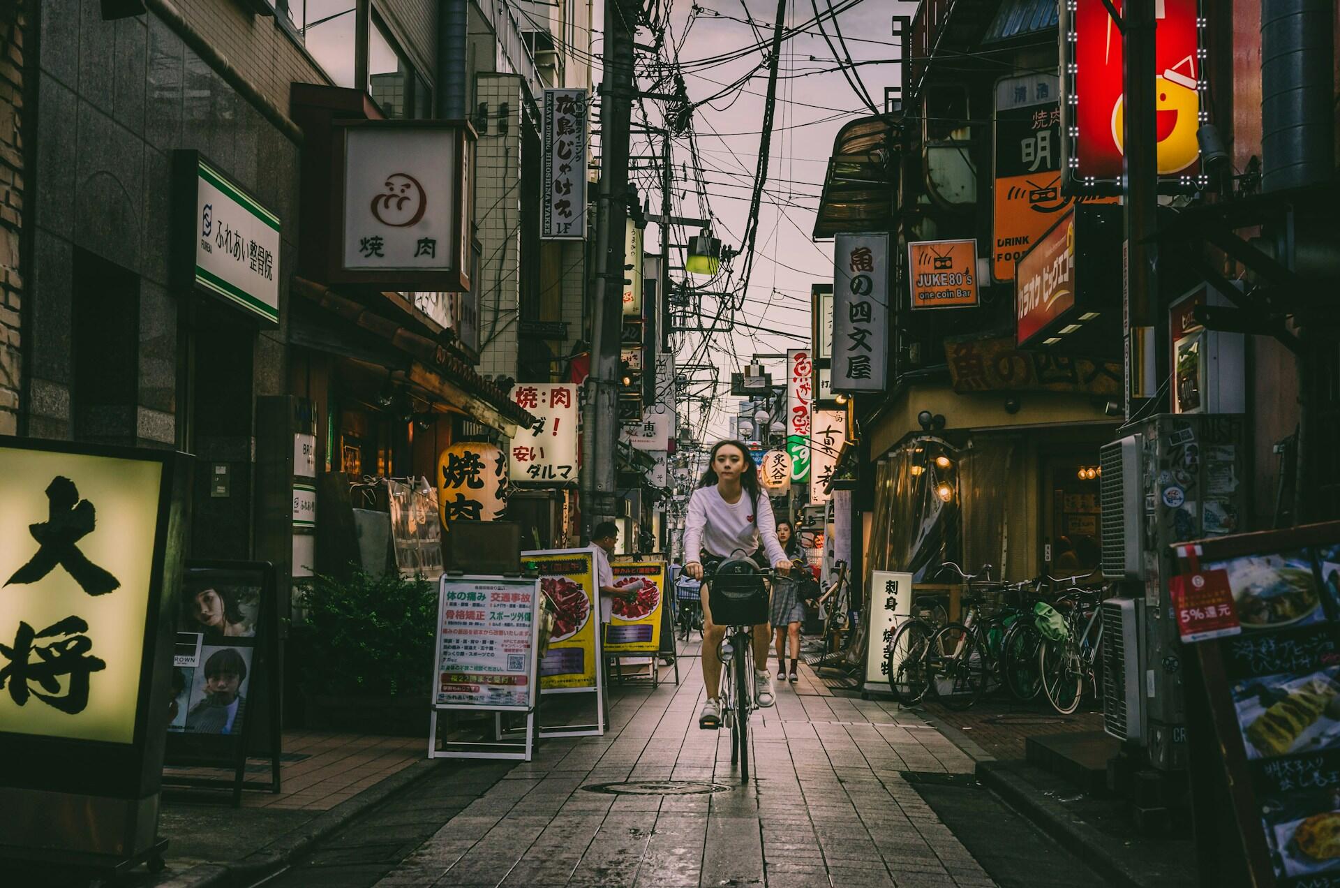 Una mujer va en bicicleta por una estrecha calle comercial de Tokio, flanqueada por letreros en japonés, restaurantes, bicicletas y faroles encendidos, bajo un cielo nublado.