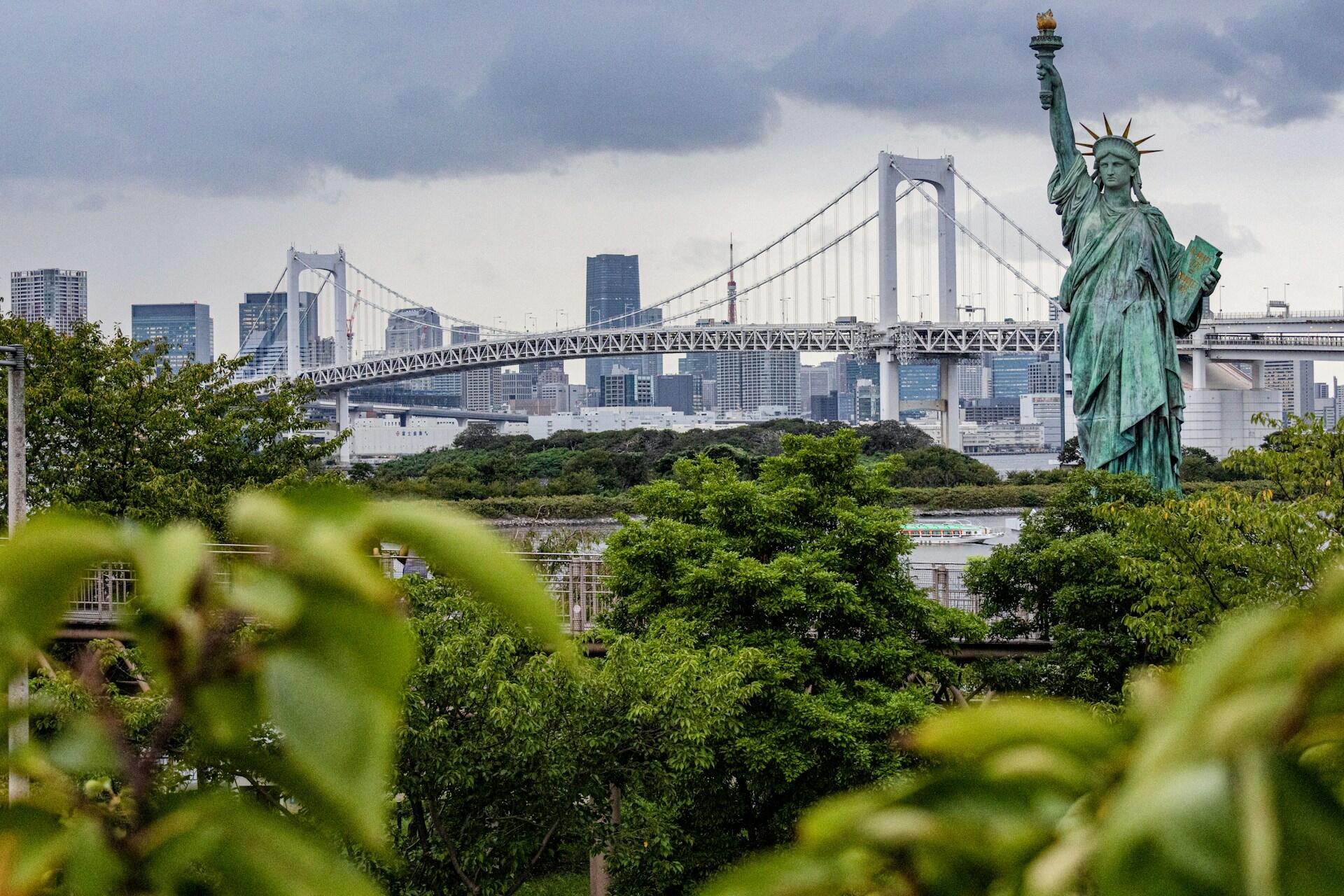 La réplica de la Estatua de la Libertad de Tokio se alza frente al Puente Rainbow y al perfil urbano, enmarcada por una exuberante vegetación bajo un cielo nublado.