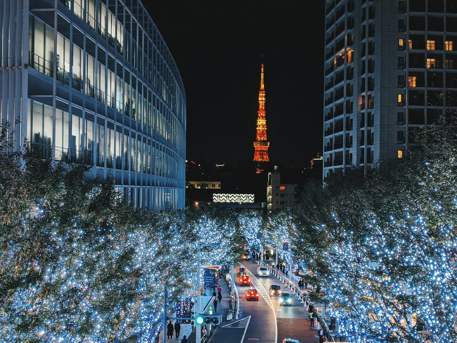 Una calle de Tokio por la noche, enmarcada por edificios modernos, con la Torre de Tokio brillando en naranja a lo lejos y los árboles resplandeciendo con luces azul-blancas.