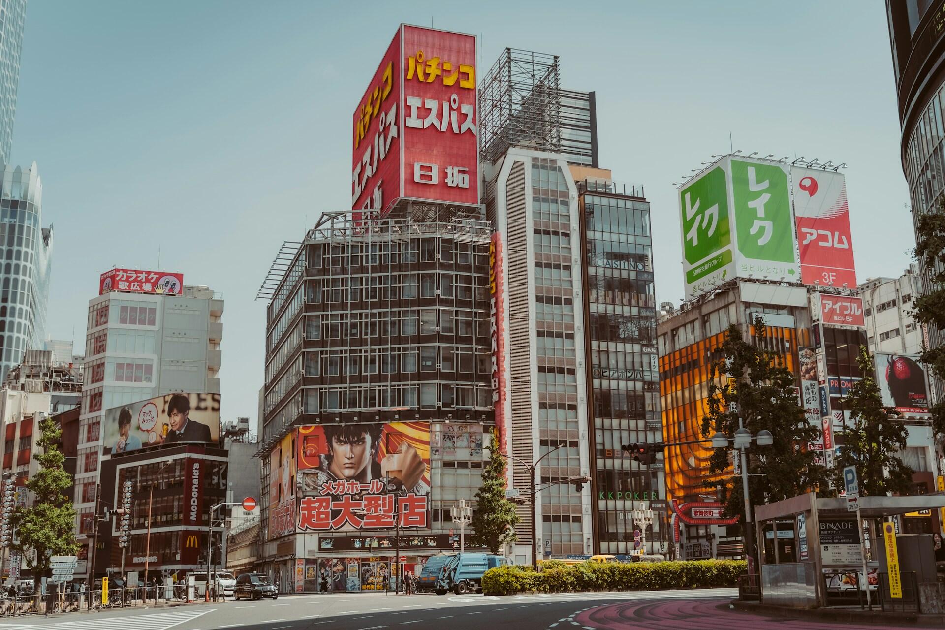 Una concurrida calle de Tokio, flanqueada por altos edificios, coloridas vallas publicitarias japonesas, tiendas, árboles y peatones, bajo un cielo azul y despejado.