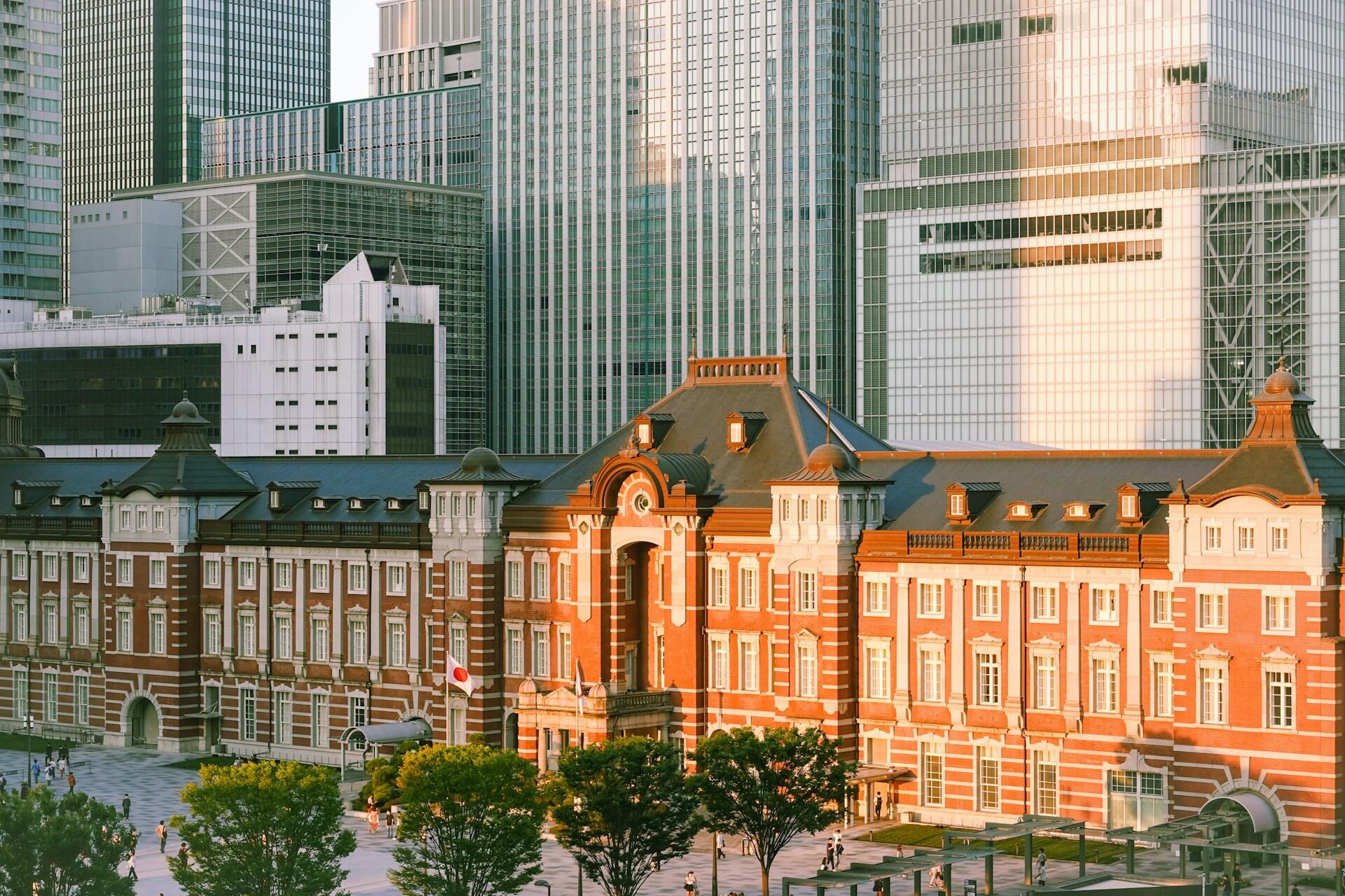 El ornamentado edificio Marunouchi de ladrillo rojo de la estación de Tokio resplandece bajo la luz del sol, enmarcado por modernos rascacielos, árboles y una animada plaza.
