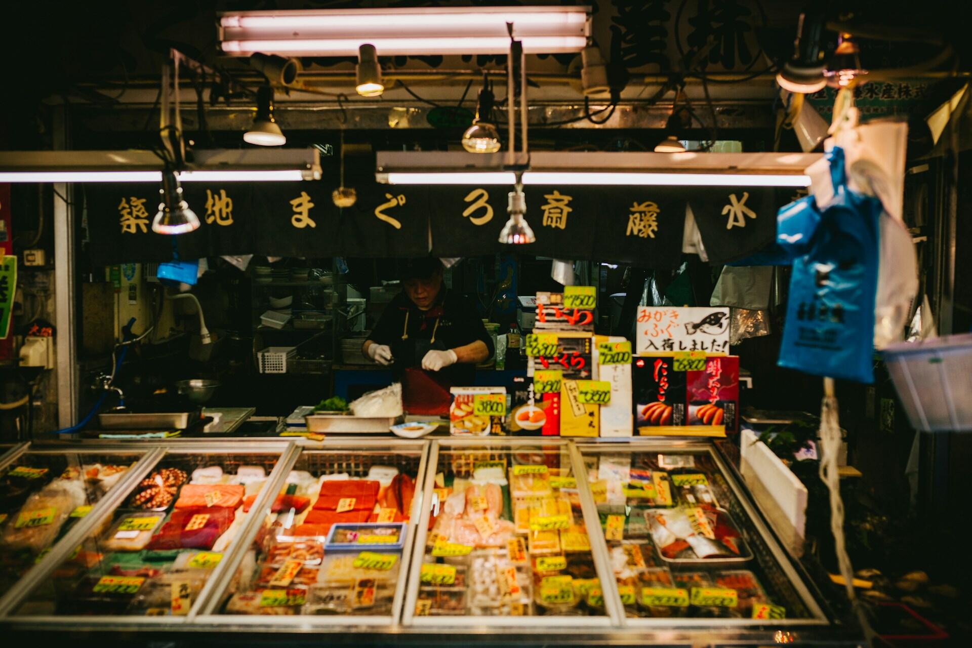 Un puesto de un mercado de marisco japonés, iluminado con una luz cálida, con un vendedor detrás de coloridas bandejas de pescado, marisco y alimentos envasados, bajo carteles escritos a mano.