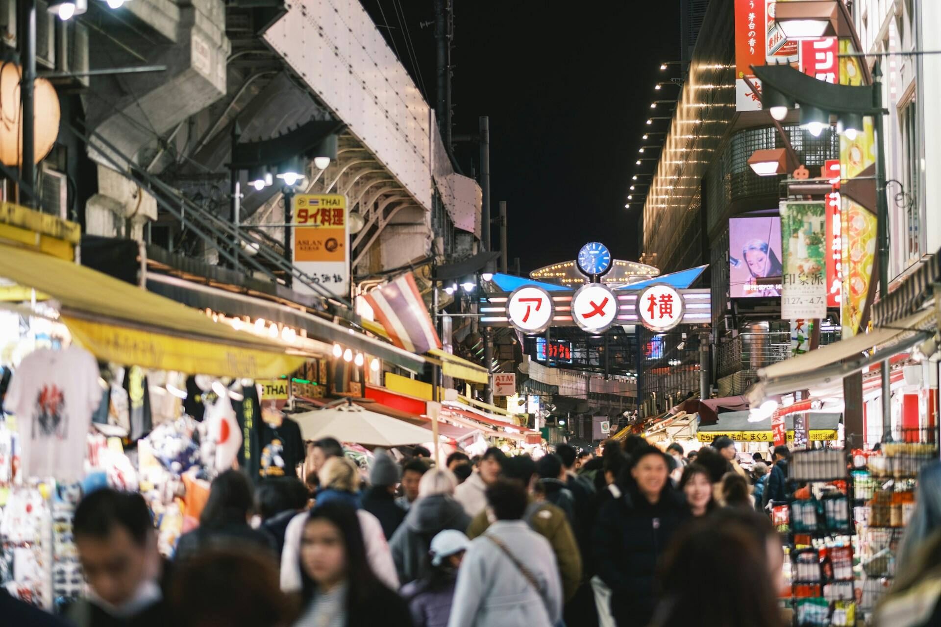 Una concurrida calle comercial japonesa por la noche, con letreros luminosos, puestos de un mercado cubierto, escaparates de colores y un animado flujo de peatones.