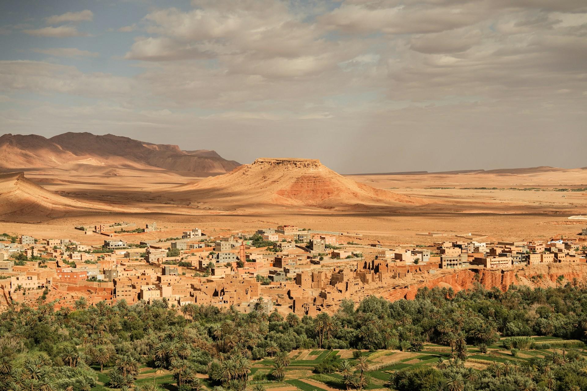 Ciudad oasis del desierto con vegetación, edificios tradicionales y una colina de cima plana bajo un cielo brumoso.