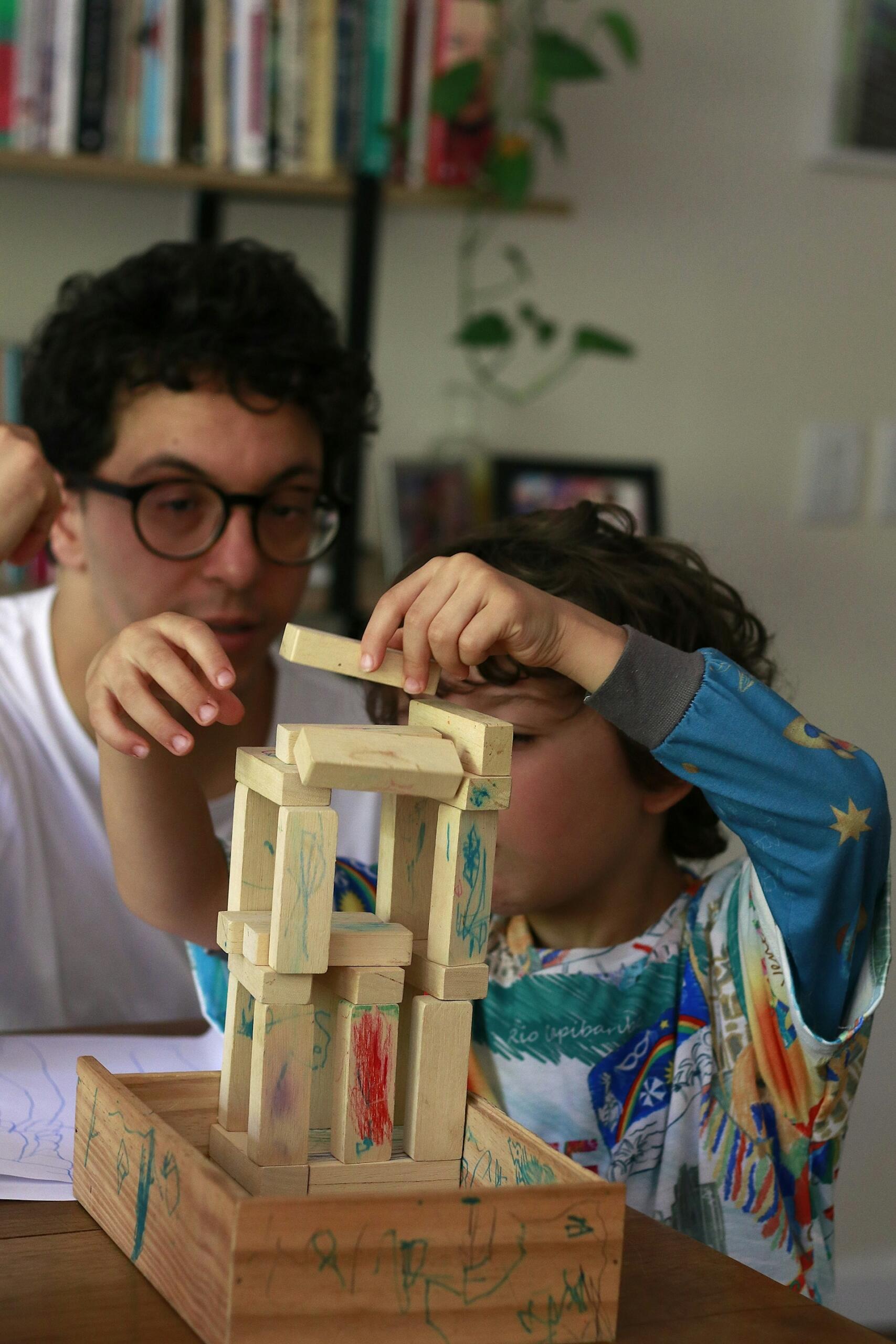 Un niño con un pijama de colores construye una torre de bloques de madera sobre una mesa, concentrado y guiado por un adulto que lleva gafas. Al fondo se ven libros y plantas.