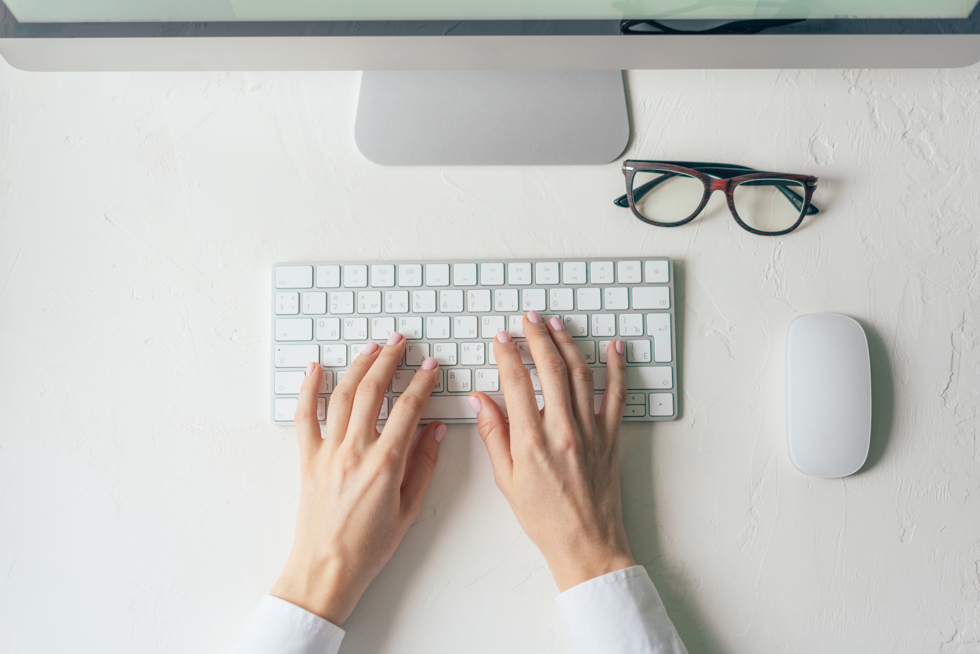 manos en taclado mac con lentes al costado derecho sobre una mesa blanca